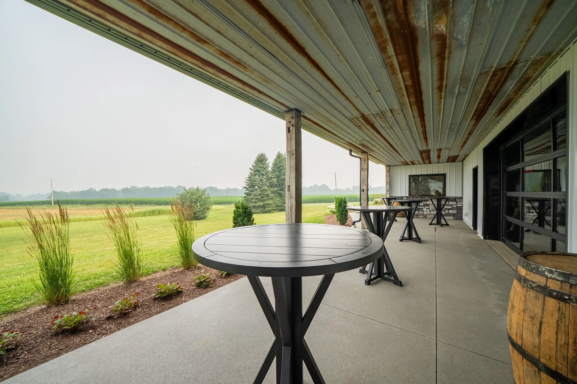 Covered outdoor patio with round tables and metal chairs, overlooking a green field with tall grass, trees, and cloudy sky.