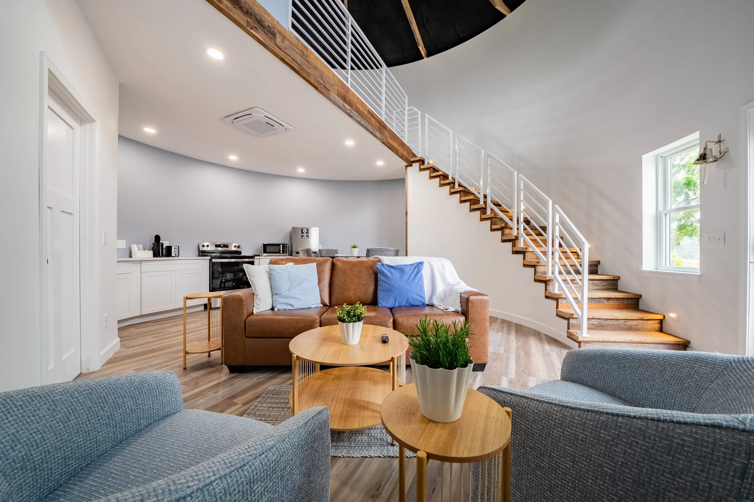 Open-concept living room with a brown leather sofa, a round wooden coffee table, blue upholstered chairs, and potted plants. In the background, a staircase with wood steps and white railing leads to the upper level, with a kitchen area visible to the left.