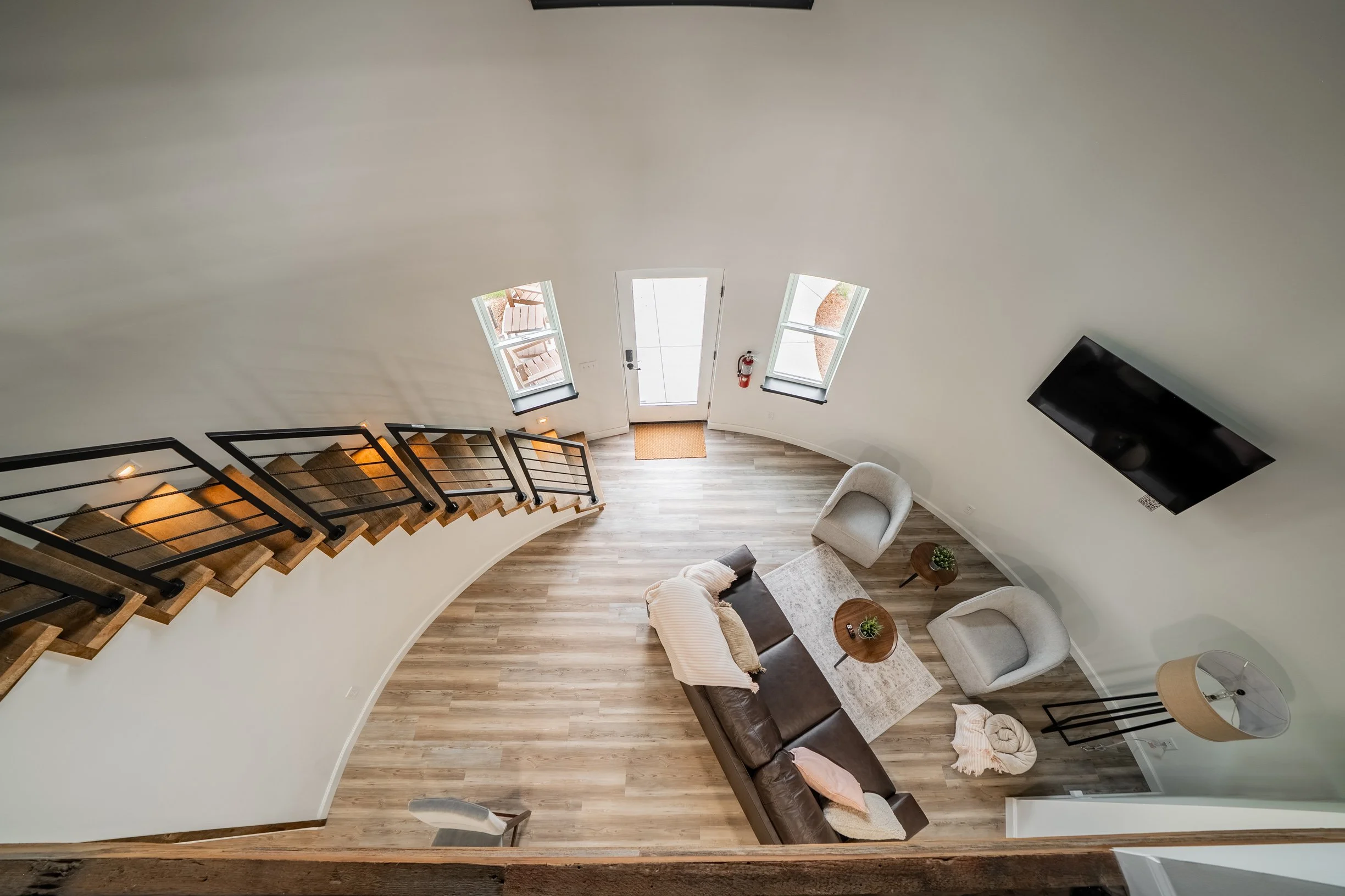 A view of a modern living room with hardwood floors, a curved wall with three windows, a staircase with black metal railing, a brown leather sofa, two white armchairs, a coffee table, a wall-mounted flat-screen TV, and decorative plants, with a door 