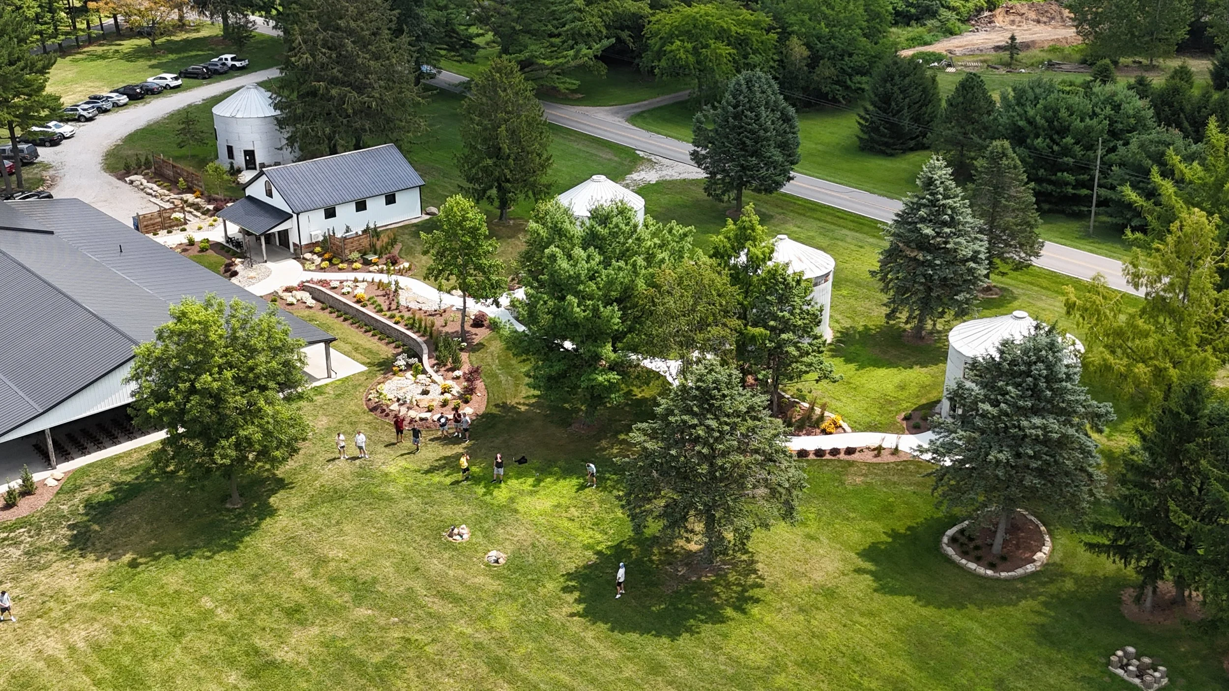 An aerial view of a green landscape with trees, a white building, silos, a parking lot, and a group of people gathered on the grass.