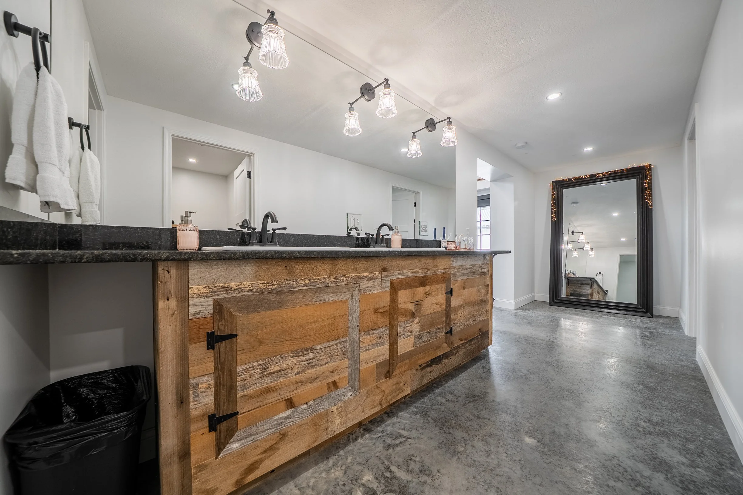 Bathroom with wooden vanity, black granite countertop, two sinks, wall-mounted towel rack with white towels, large framed mirror with decorative lights, and ceiling lights.