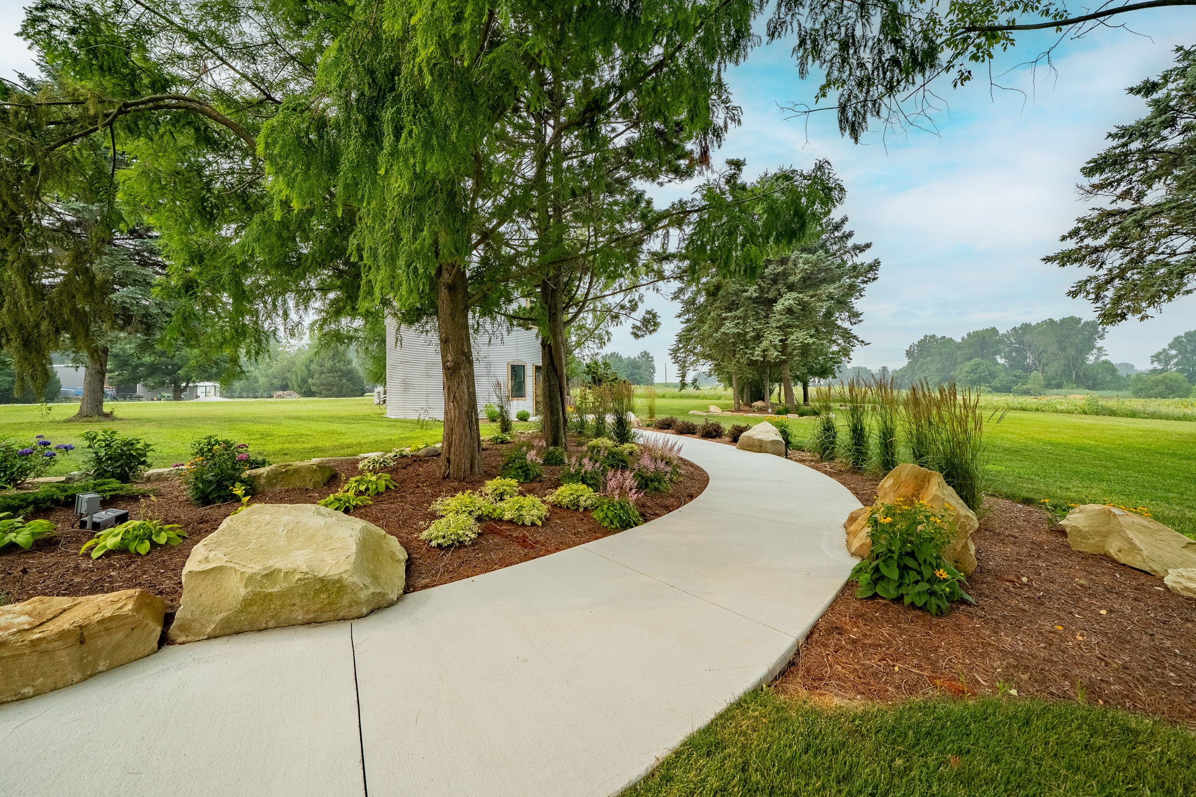 A landscaped garden with a paved walkway, large rocks, lush green trees, and various plants and flowers, under a partly cloudy sky.