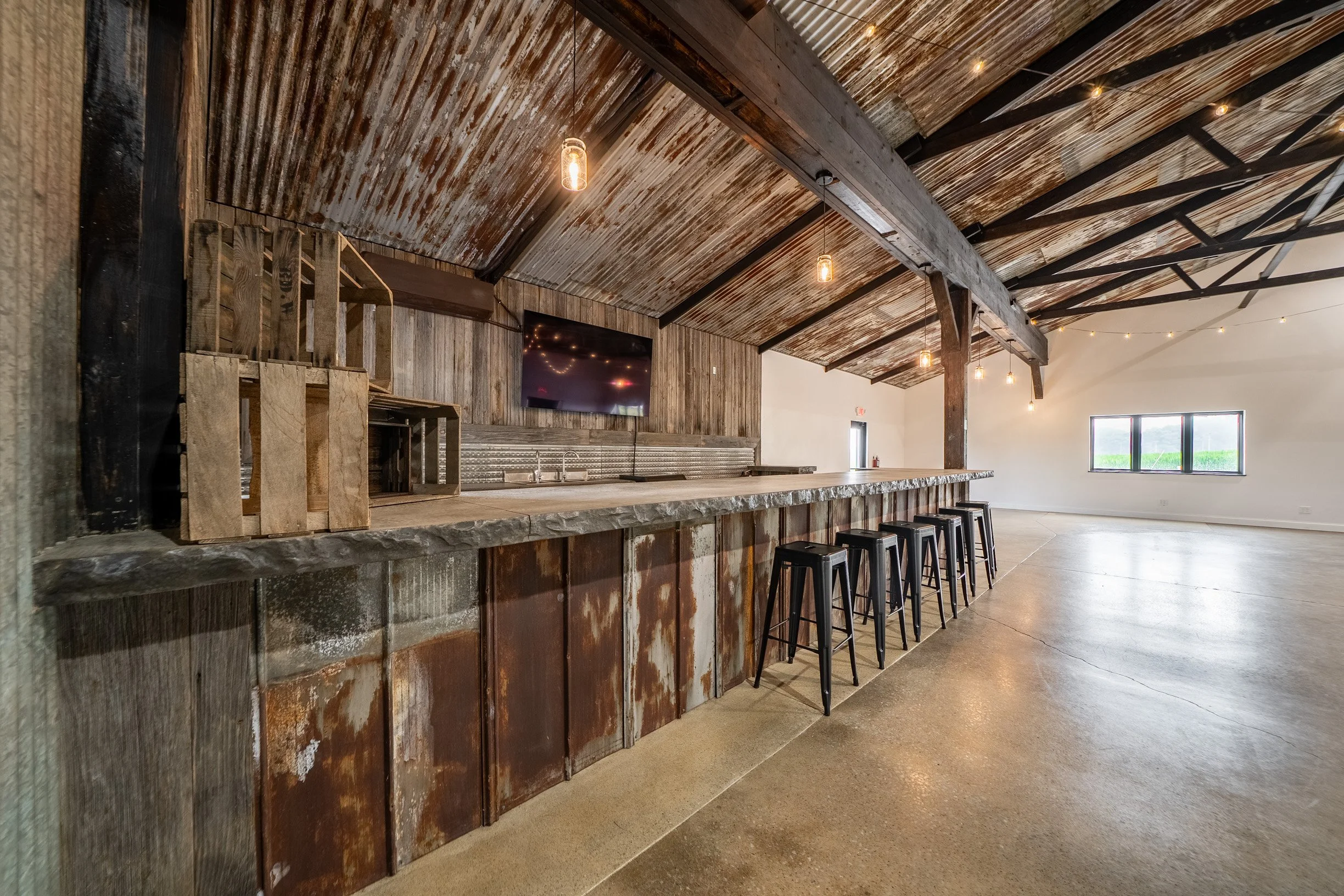 Interior of a rustic barn-style space with a high, curved, wood-paneled ceiling and string lights. There is a bar with a concrete countertop, simple black metal stools, and a flat-screen TV on the wall.