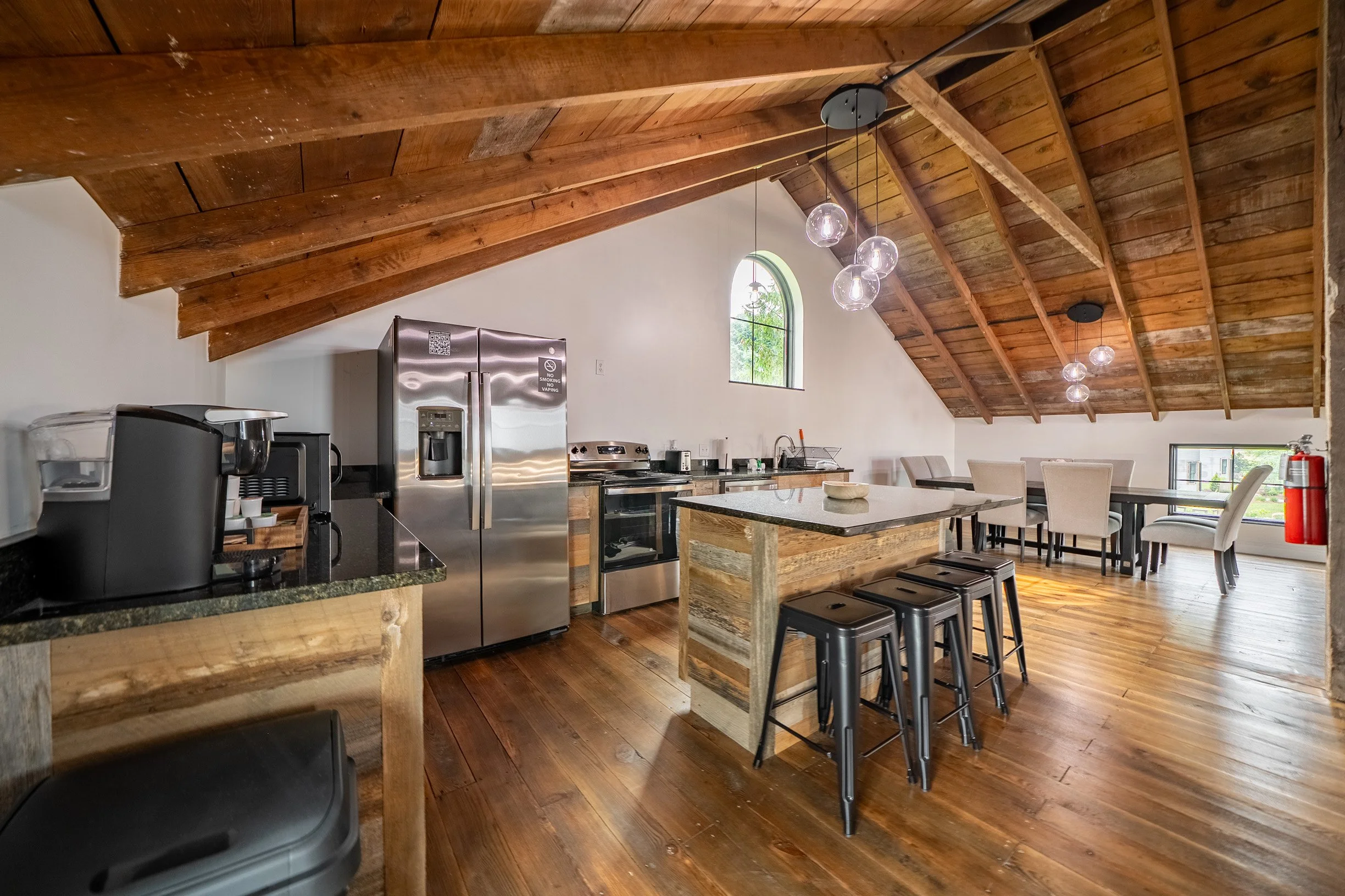 A spacious kitchen with a high wooden vaulted ceiling, hanging globe pendant lights, a stainless steel refrigerator, kitchen island, white chairs around a dining table, and a window with greenery outside.