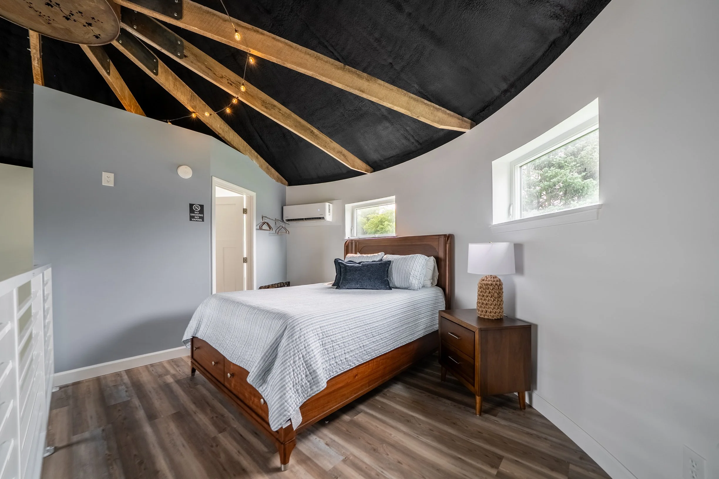 A bedroom with a wooden bed frame, gray and white bedding, and a dark textured ceiling with wooden beams and string lights. There is a nightstand with a lamp and window showing trees outside.