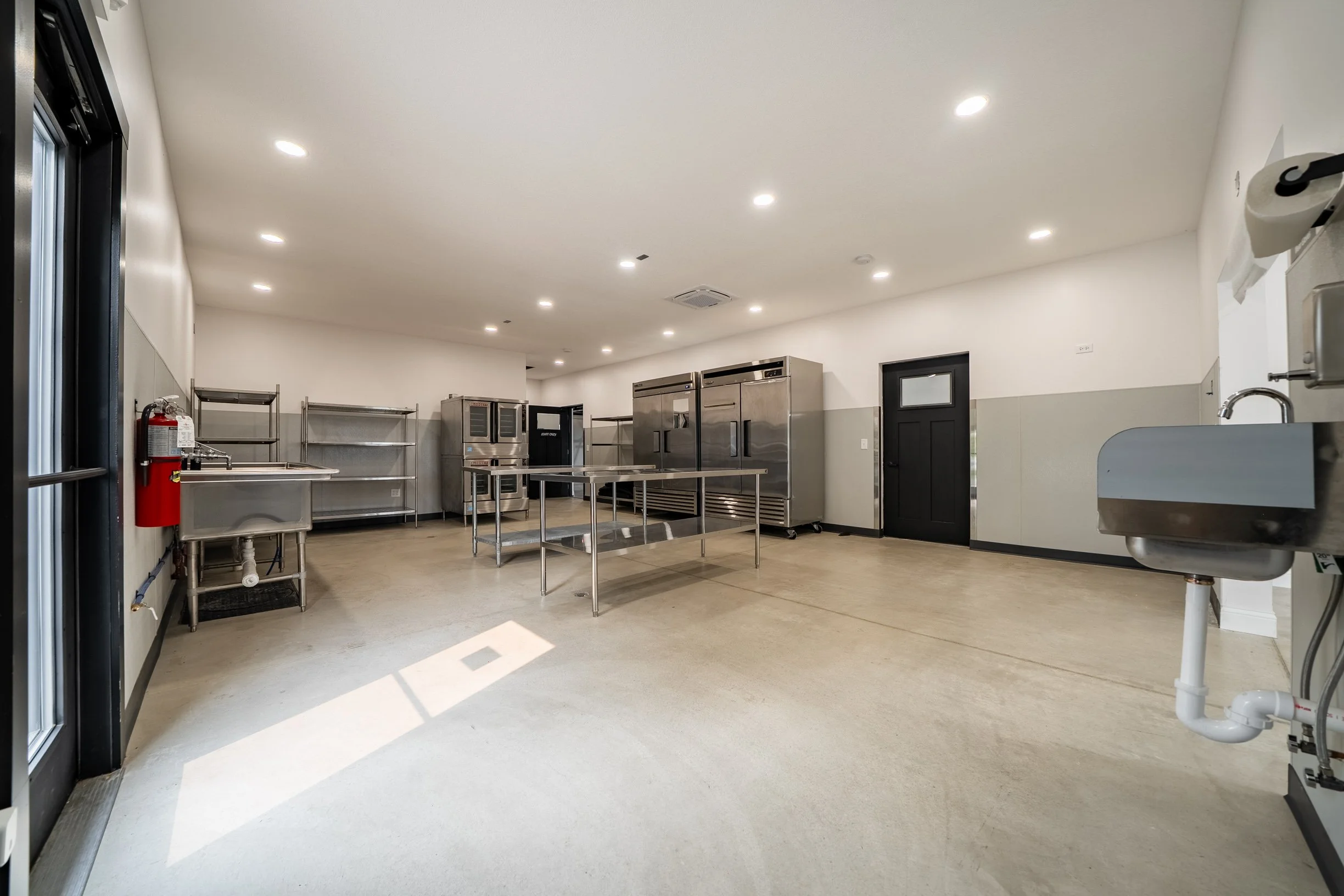Empty commercial kitchen with stainless steel appliances, metal shelving, a utility sink, and a fire extinguisher, illuminated by recessed ceiling lights.