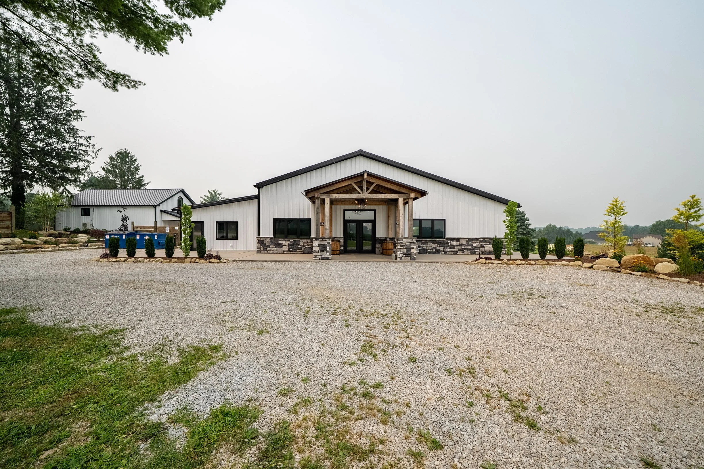 Modern white building with stone accents and a wooden porch, set on a gravel lot with trees and greenery around.