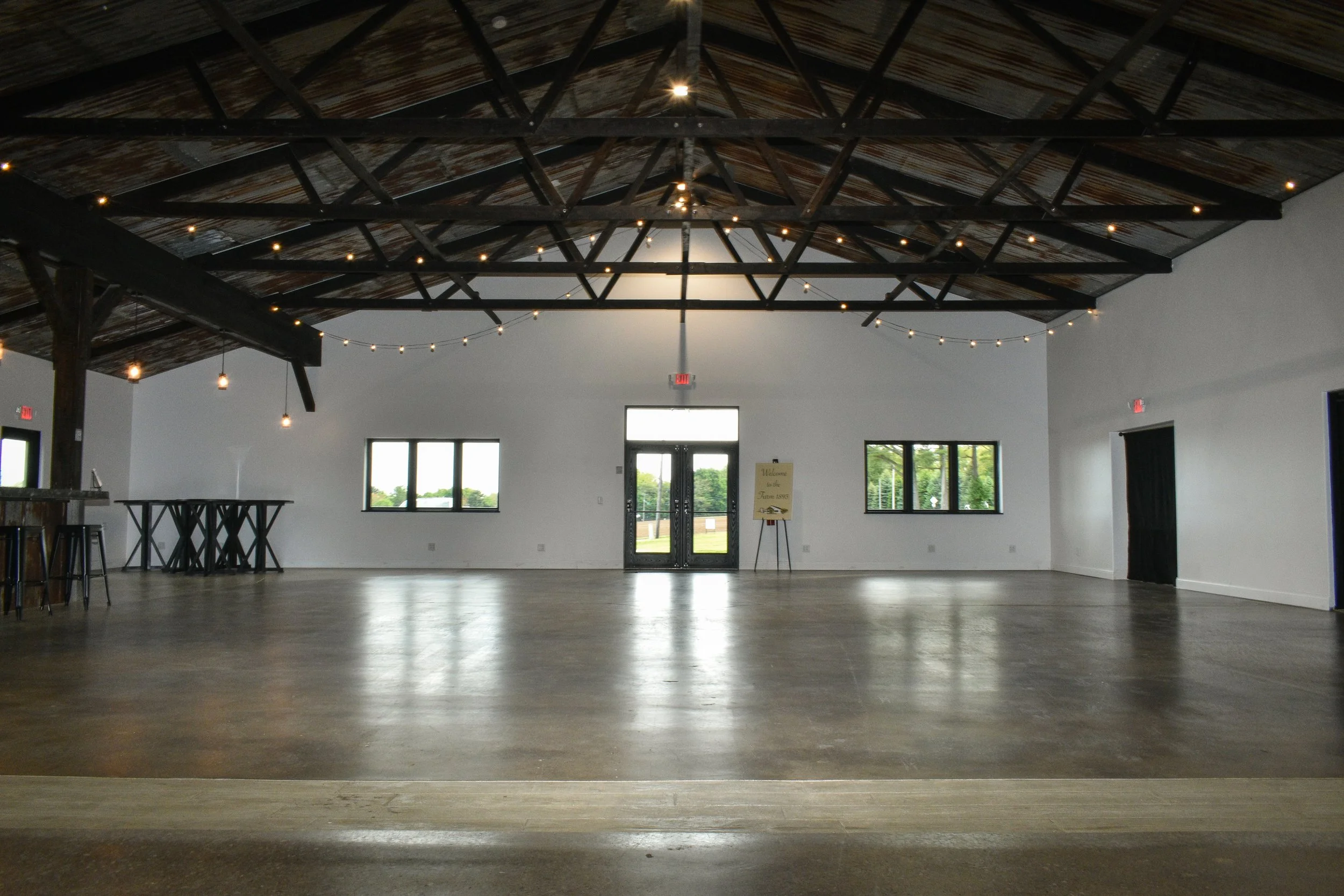 Empty indoor event space with polished wood floor, black metal ceiling beams with string lights, and large windows with a view of trees outside.