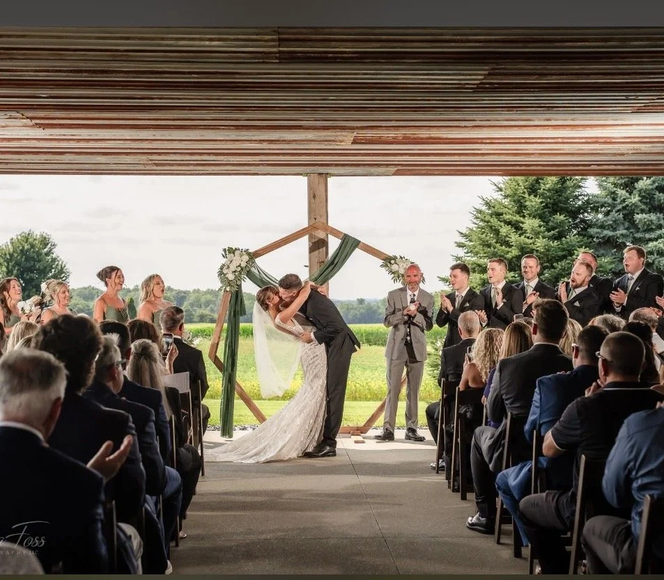 A wedding ceremony with the bride and groom kissing under a wooden frame decorated with flowers and green fabric, surrounded by guests and the outdoor landscape.