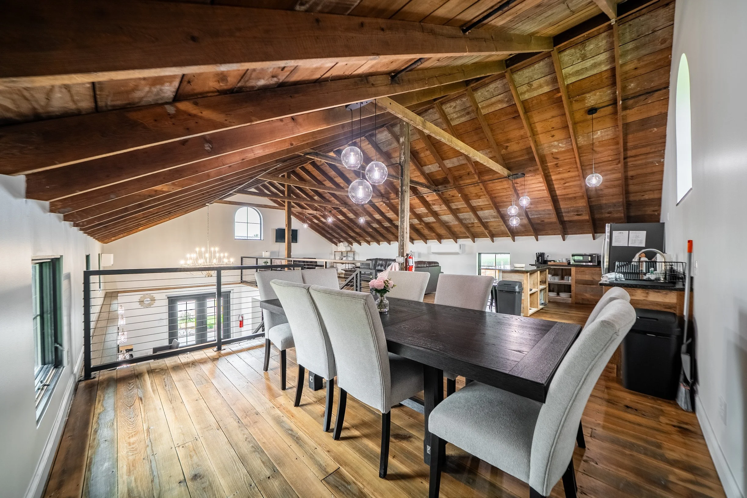 A spacious room with a high vaulted wooden ceiling, hanging light fixtures, a large dining table with chairs, and a kitchen area in the background.