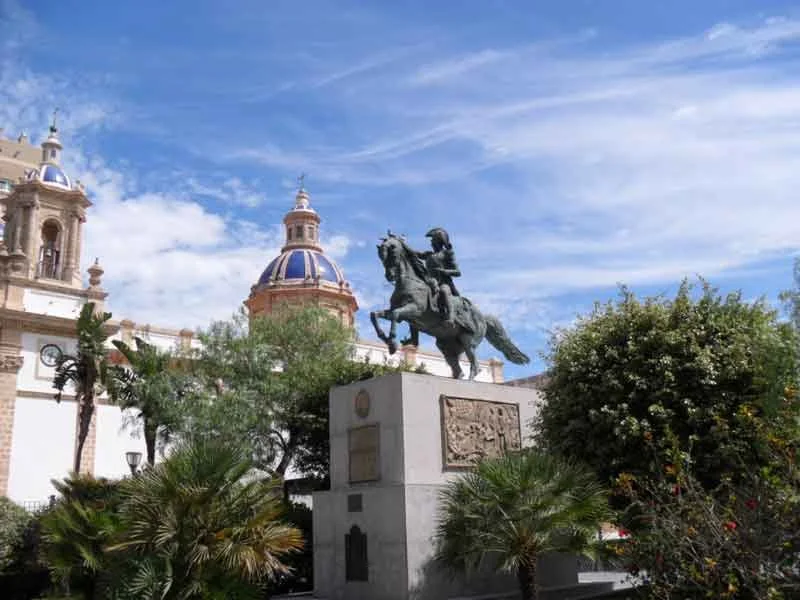 Statue of a man on horseback in front of a building with domes, surrounded by trees and bushes, under a blue sky with clouds.