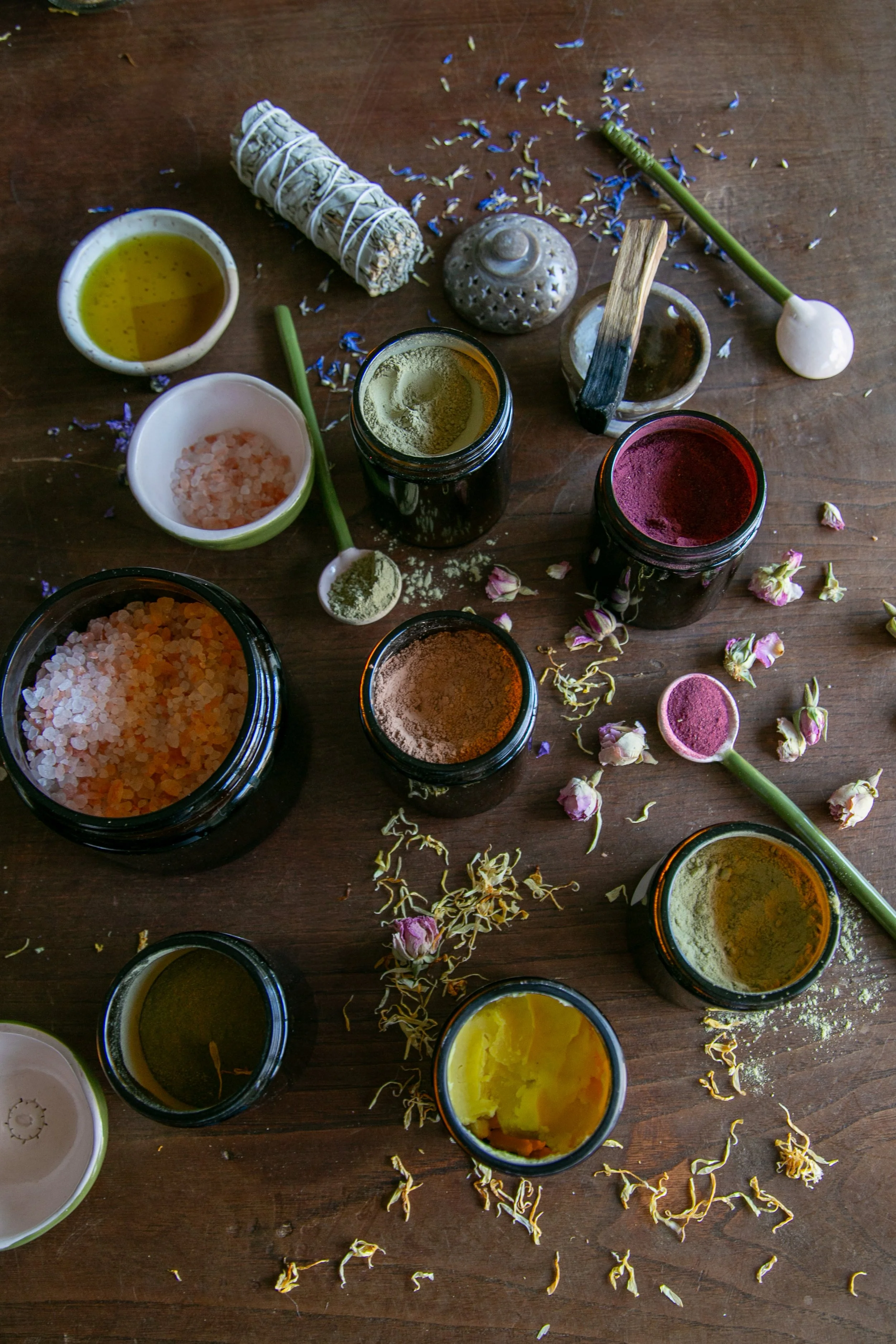 Collection de herbes, de poudres colorées, de sels et d'huiles essentielles sur une table en bois, utilisés pour des rituels ou des soins.