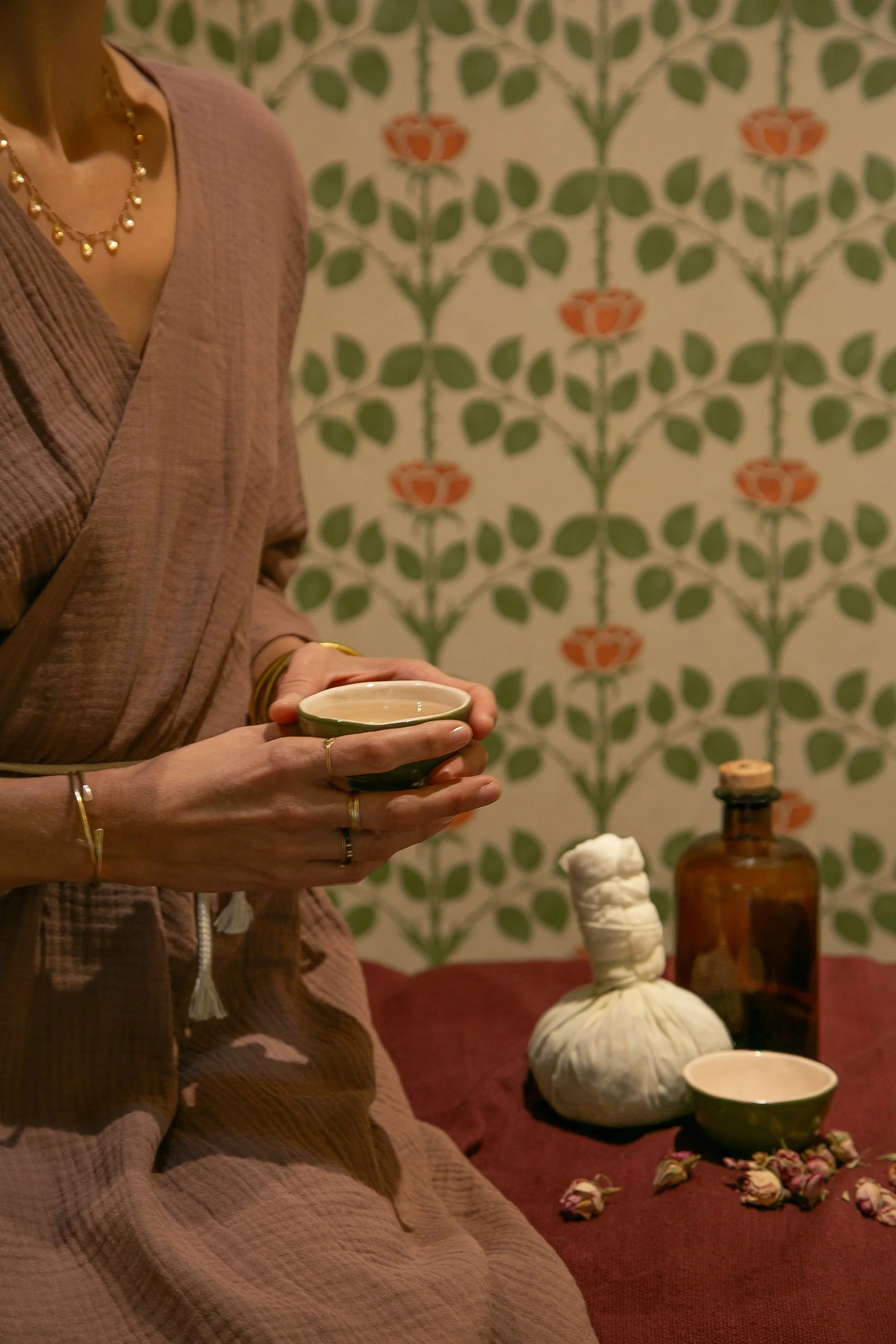Une femme assise, tenant un bol en céramique verte, portant des bijoux dorés, devant un mur avec un motif floral. Sur la table devant elle, il y a une bouteille en verre ambré, une poupée en tissu beige, une tasse en céramique verte et quelques fleurs séchées.
