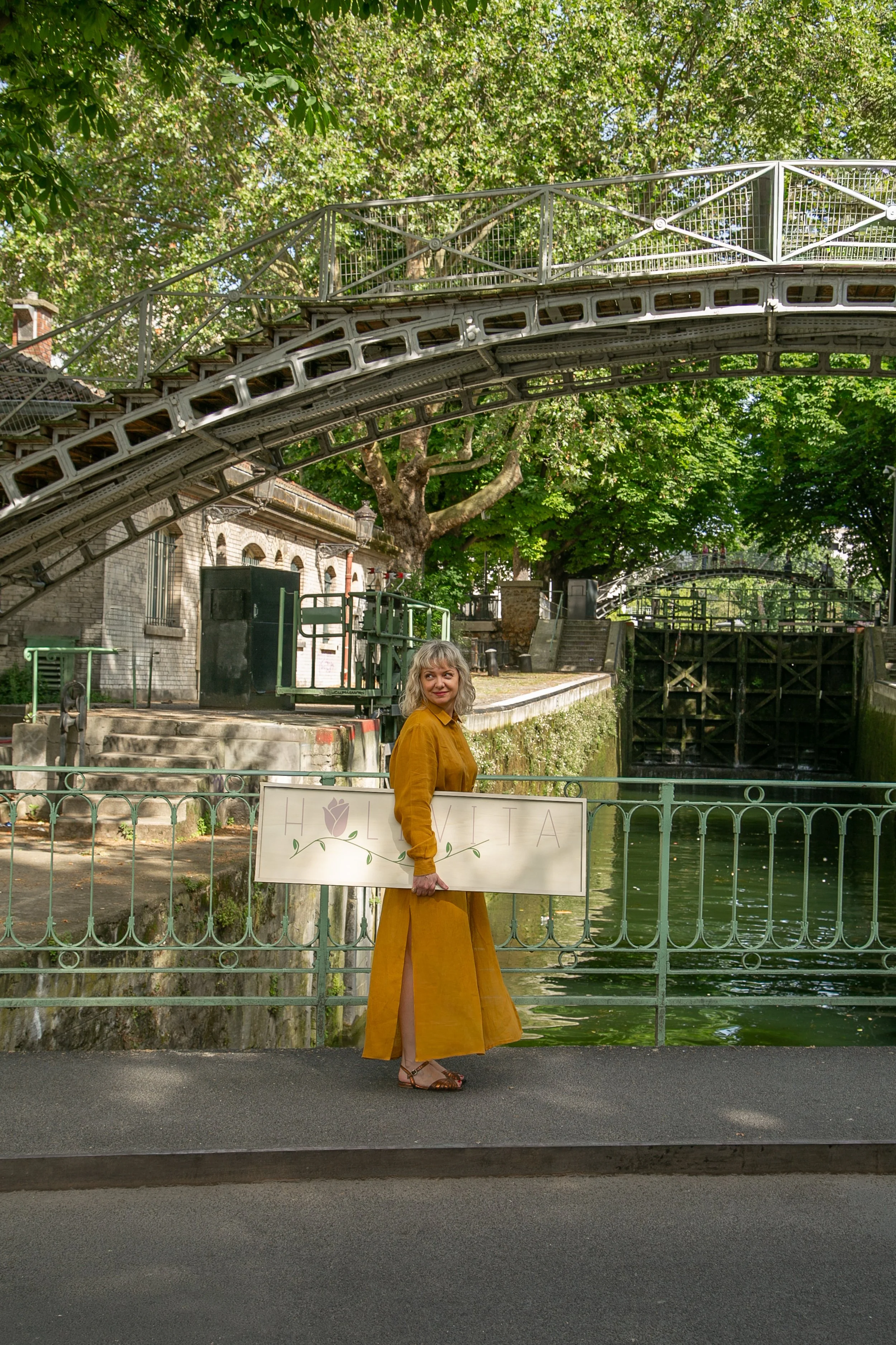 Femme portant une robe longue jaune, tenant une pancarte 'HOLIVITA'. Elle se tient près du Canal St Martin, avec un pont en métal au-dessus et de nombreux arbres verts en arrière-plan.