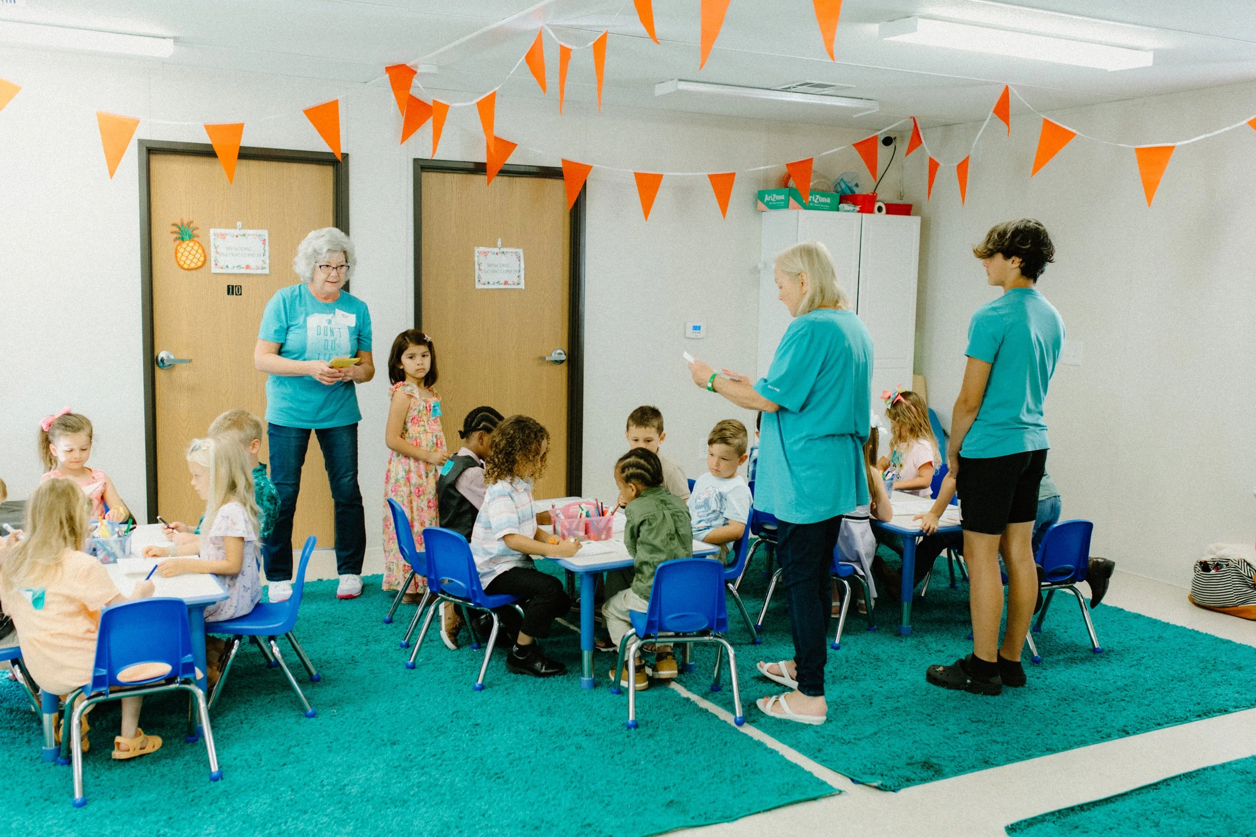 Children sitting at tables in a classroom with teachers standing nearby, orange bunting hanging from the ceiling, and pineapple and welcome signs on the doors.