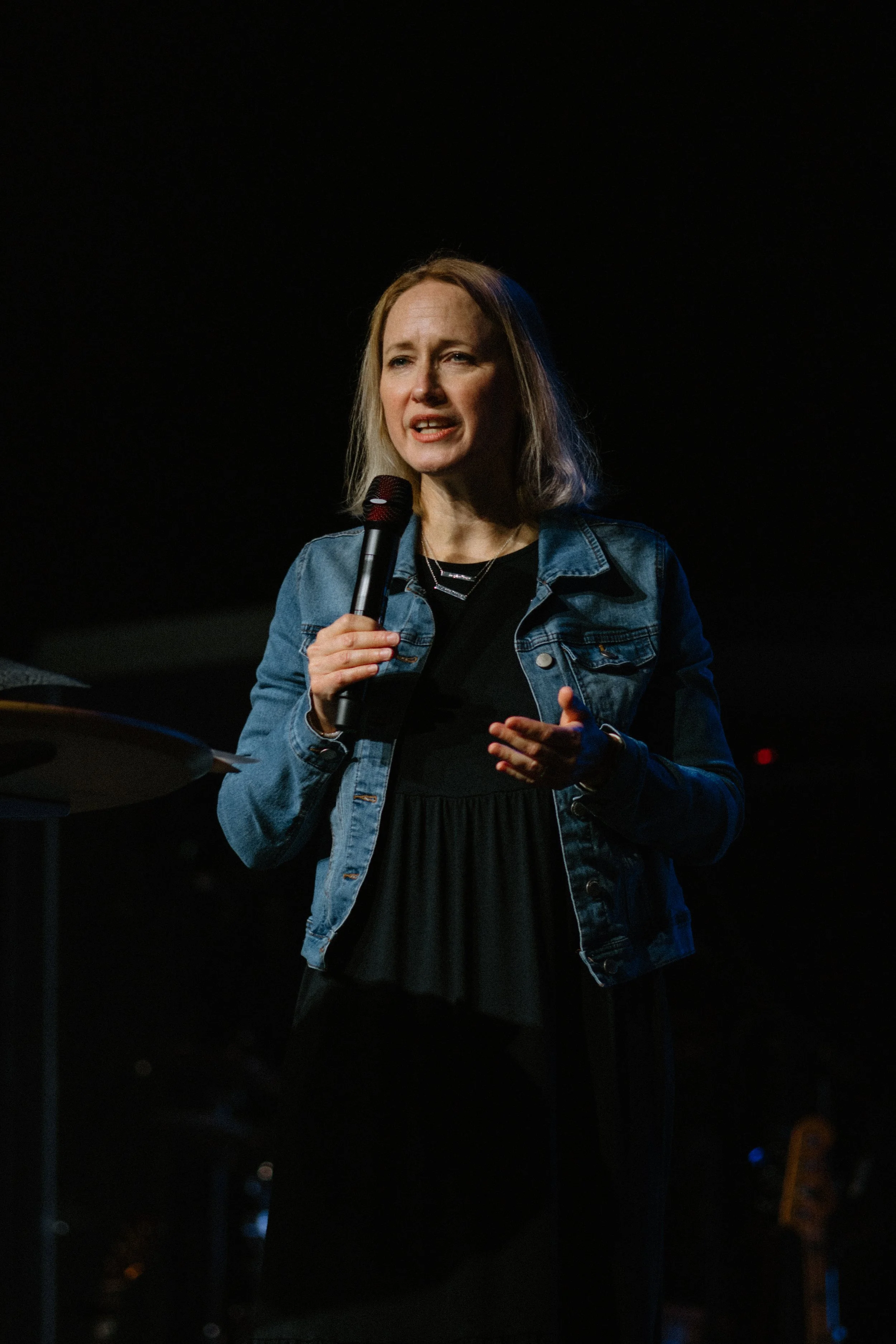 A woman with shoulder-length blond hair, wearing a denim jacket and a black dress, is speaking into a microphone on stage with a dark background.
