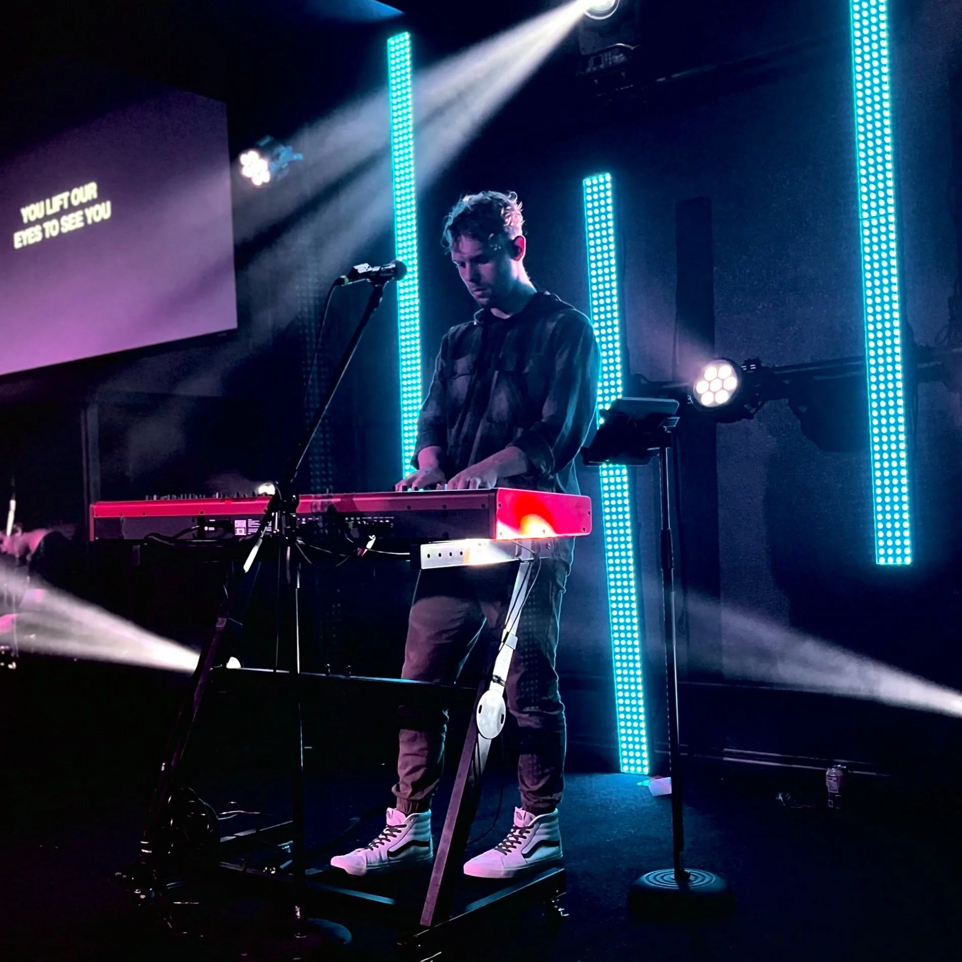 A young male musician performing on stage with electronic keyboard, illuminated by blue vertical LED lights, with a screen displaying lyrics in the background.