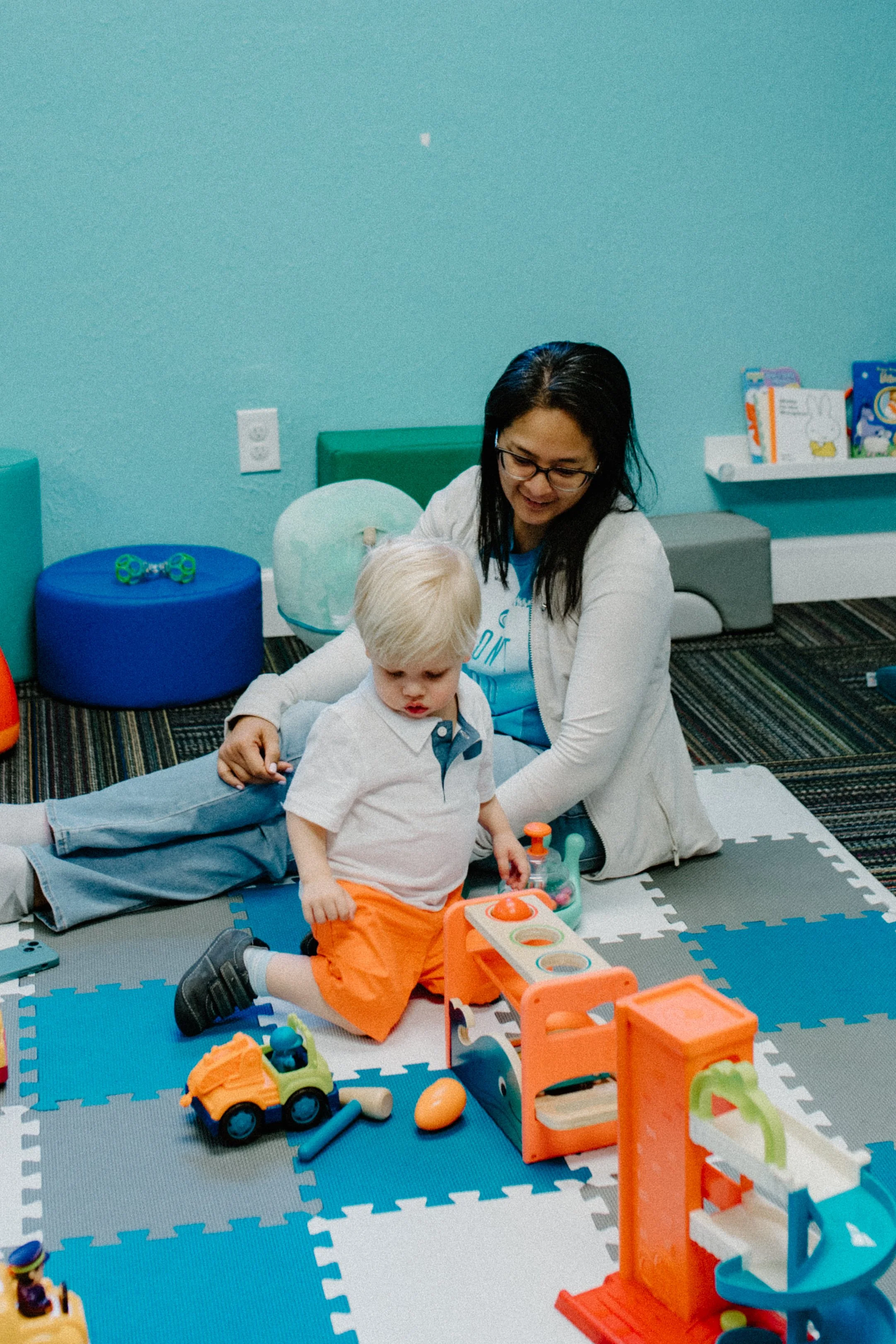 A woman with glasses and a white jacket playing with a young boy in a colorful indoor play area. The boy has blonde hair and is wearing a white shirt and orange shorts. There are toys around them on a foam mat.
