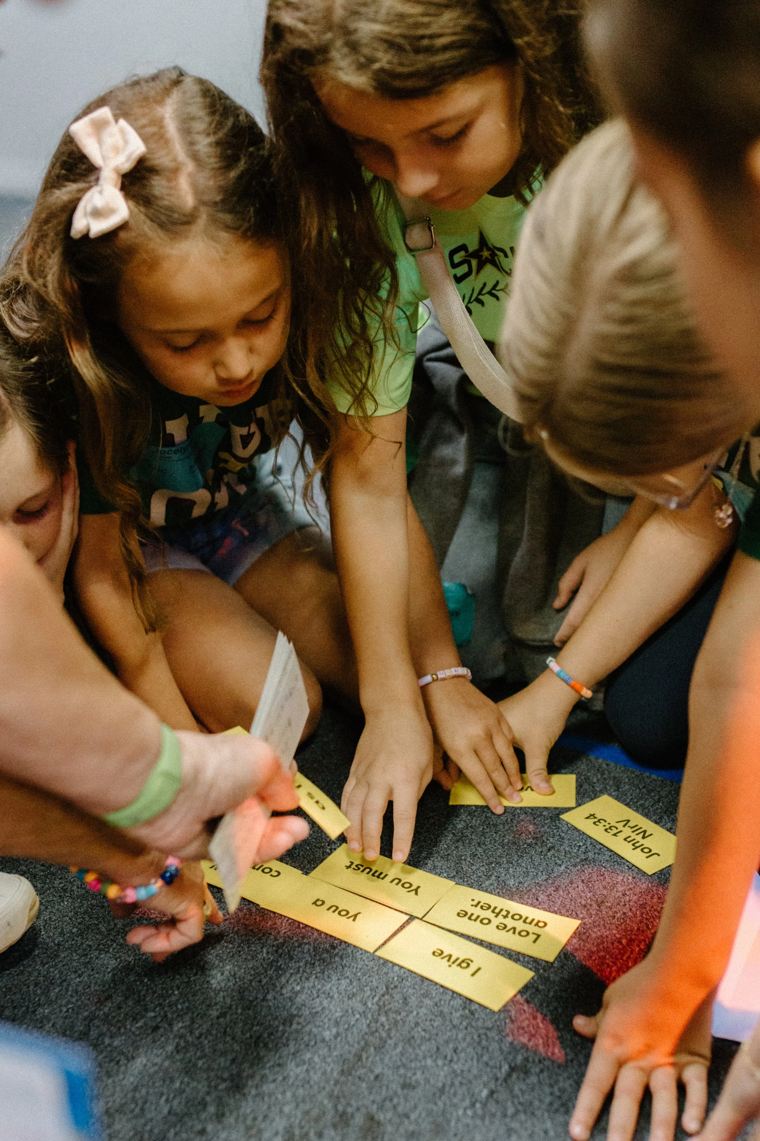 Children sitting on benches facing a stage with a person speaking into a microphone and a large screen displaying text in a dark room with colorful lighting.