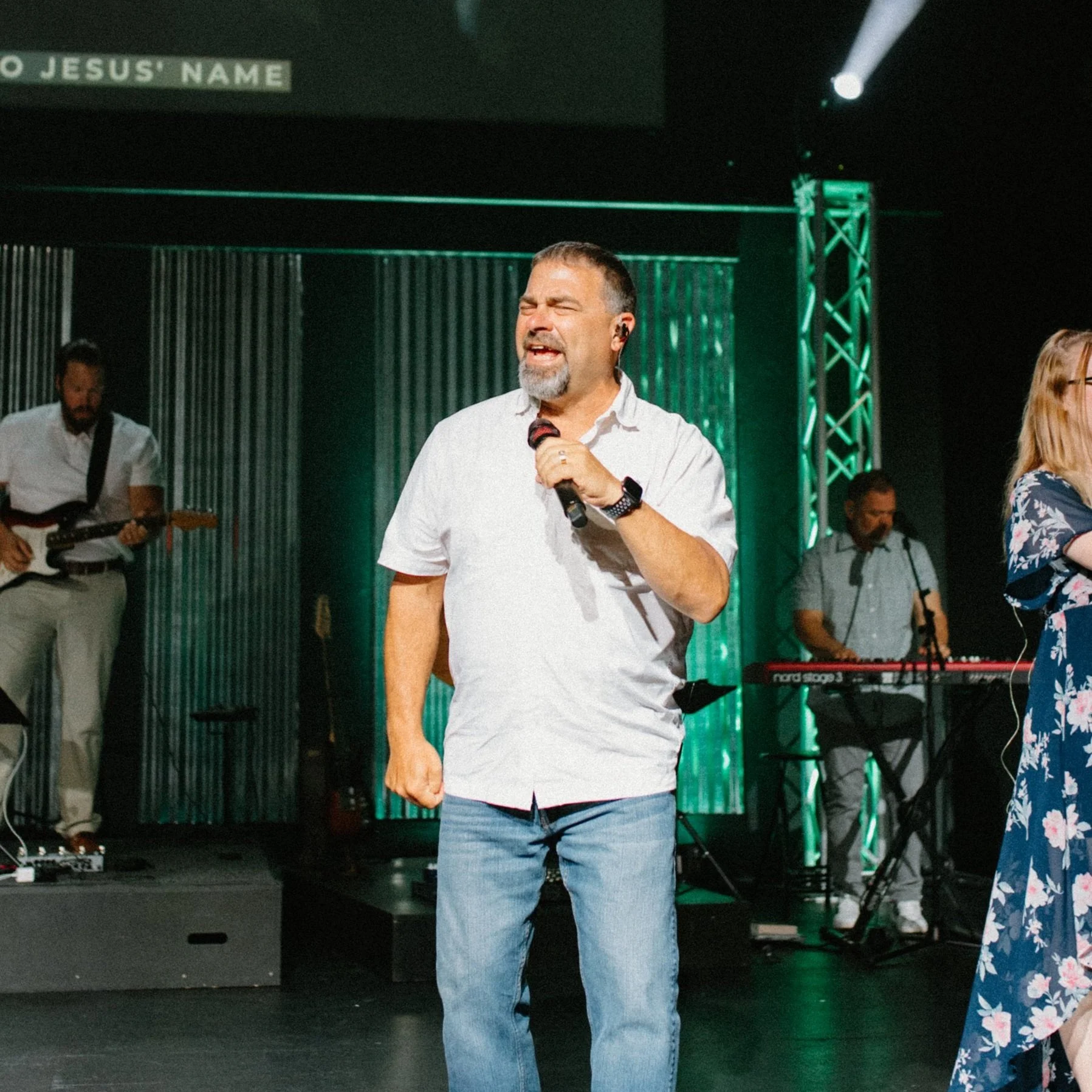 A man singing on stage with a microphone, wearing a white shirt and jeans, with a band playing instruments in the background.