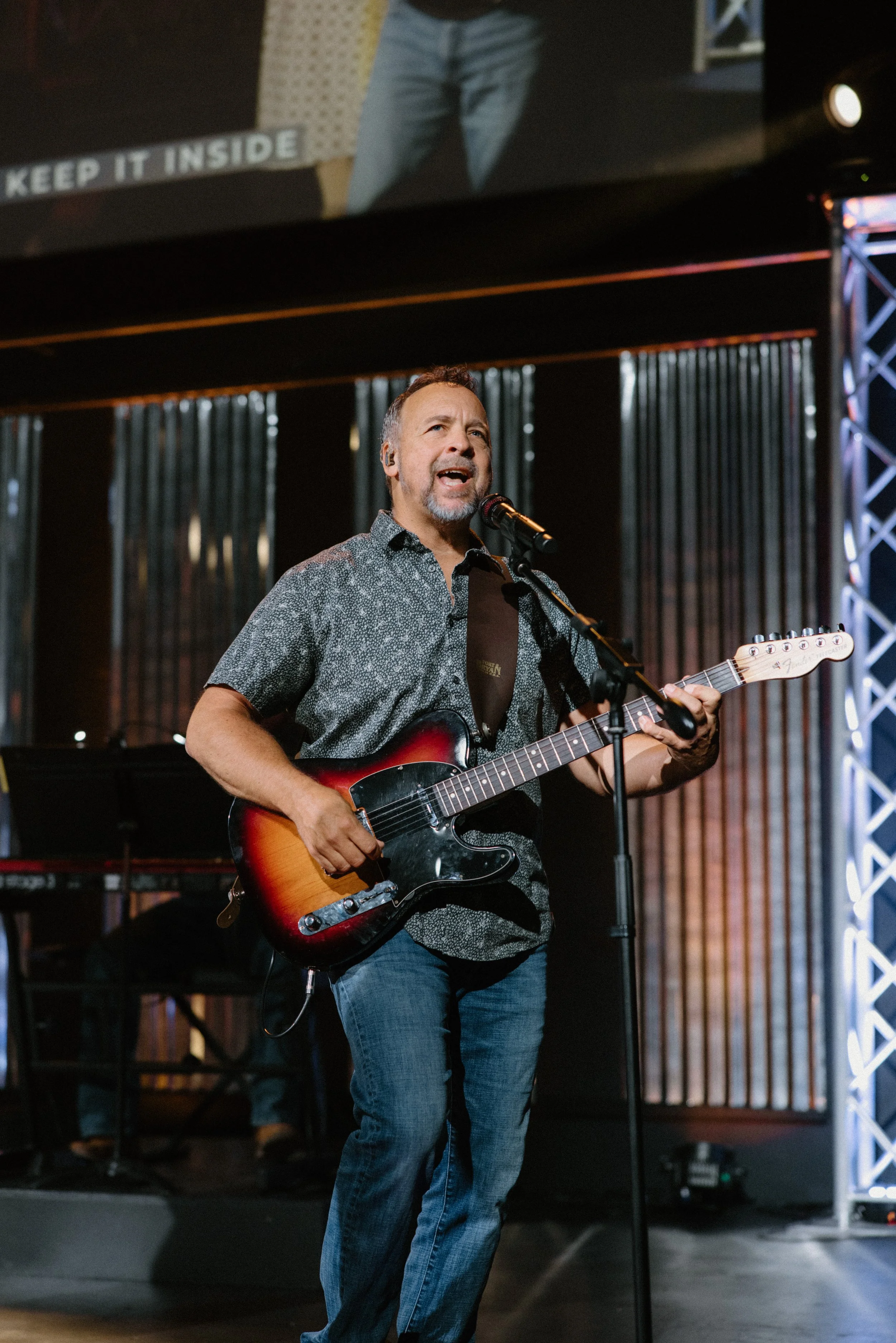 A man singing into a microphone and playing an electric guitar on a stage with a dark background and lighting.