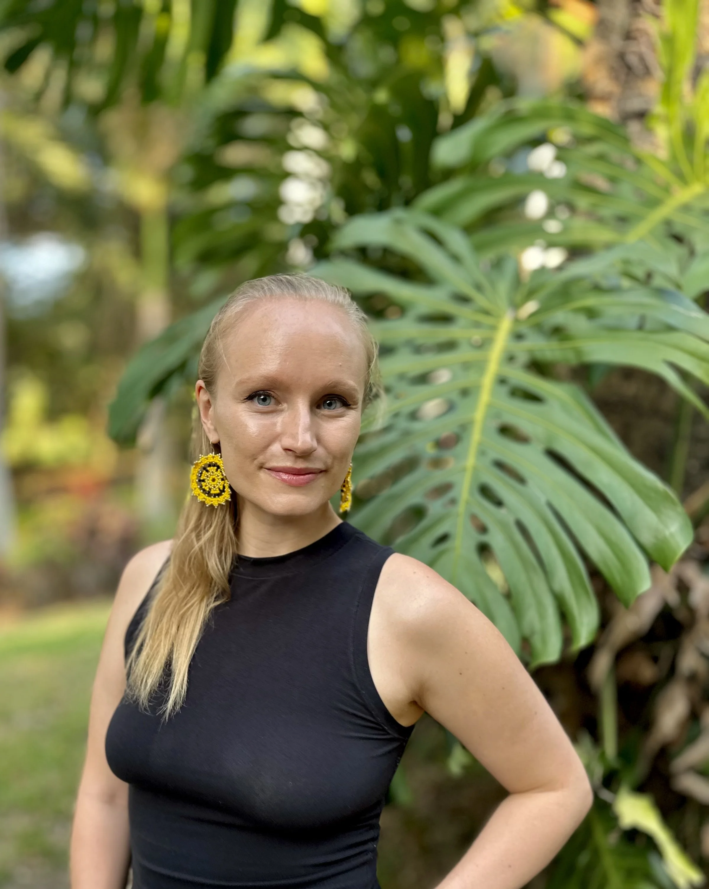 Woman in black sleeveless top and yellow earrings standing in front of large green leaves outdoors.