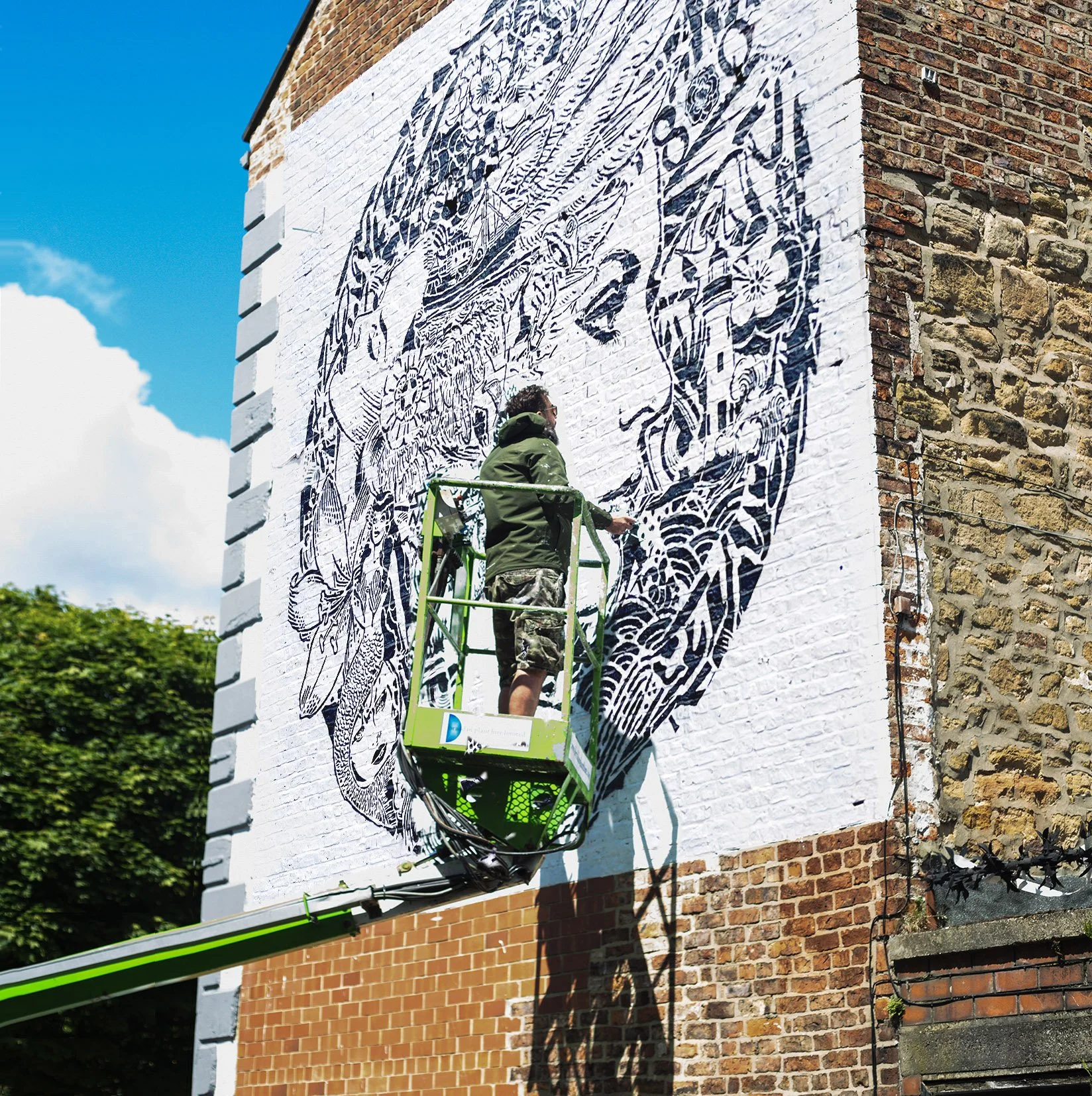 Artist Prefab77 on a cherry picker working on a black and white mural showing an intricate design of a woman's face on a whitewashed brick wall gable end