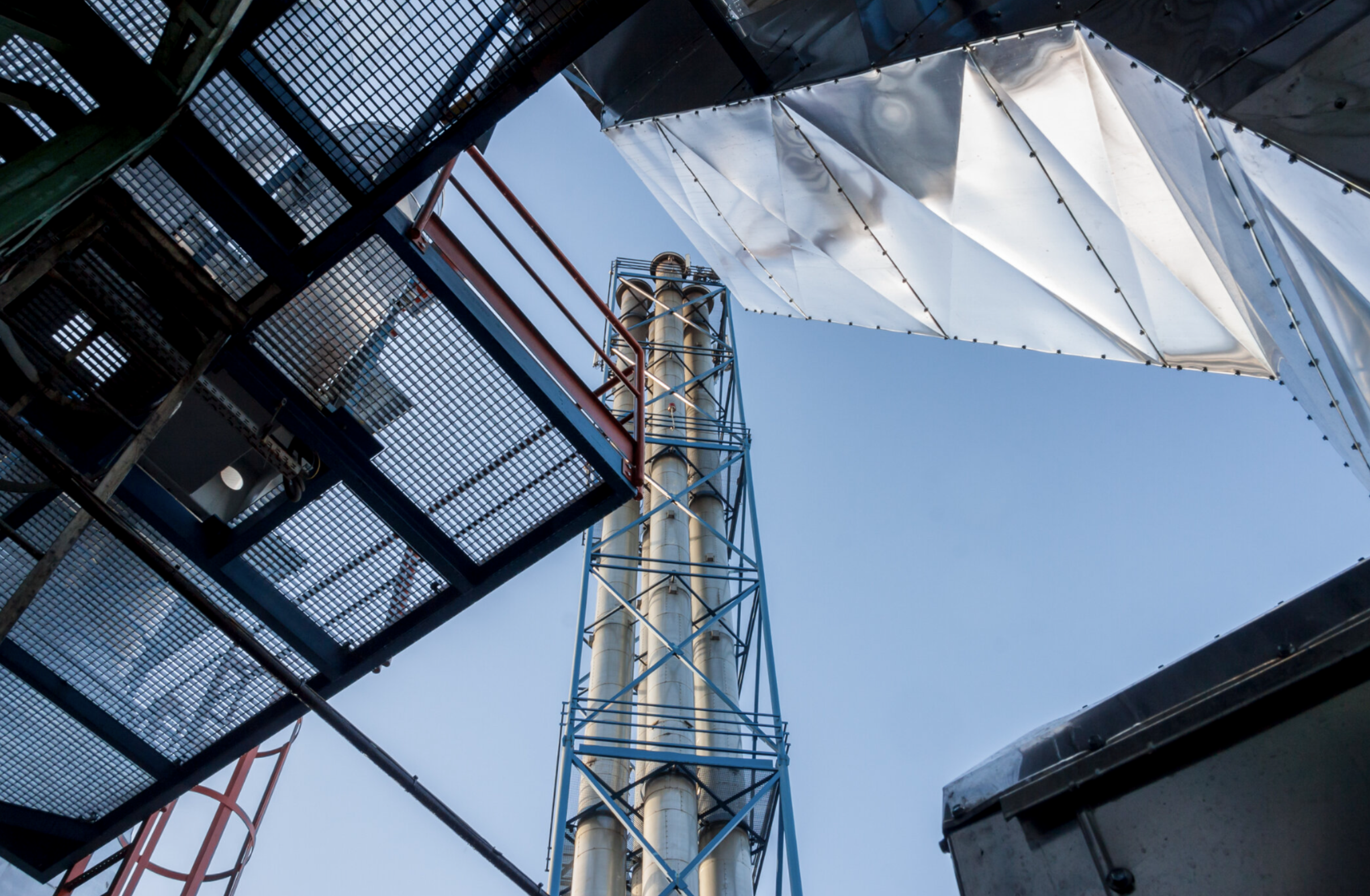 Veduta dal basso di una torre industriale circondata da strutture in metallo e reti, con il cielo azzurro sullo sfondo.