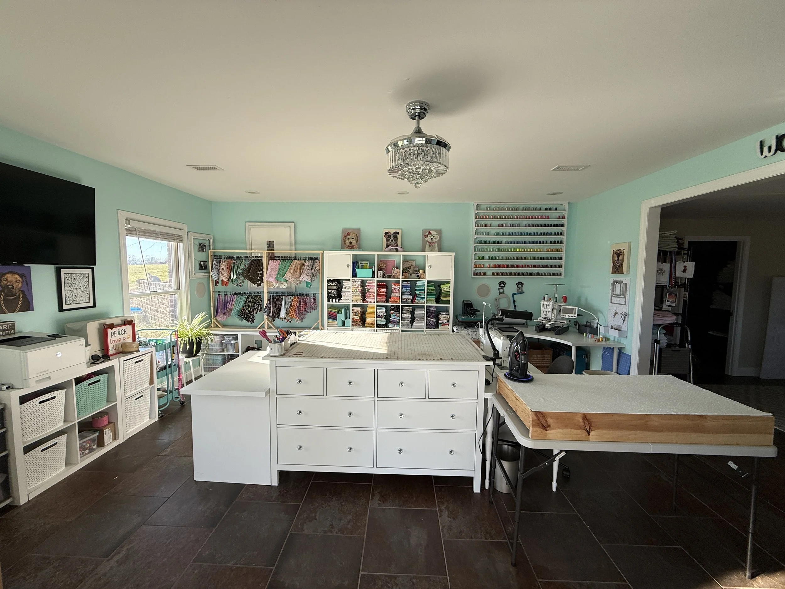 A craft room with pastel mint green walls, featuring a large central white storage island, a wall-mounted flat-screen TV, a window with sunlight, colorful fabric swatches, organized shelves with craft supplies, a work desk with sewing equipment, and a chandelier hanging from the ceiling.
