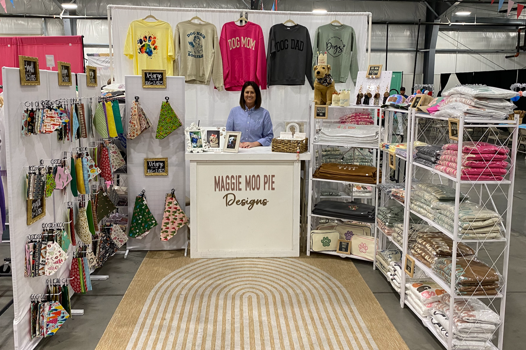A woman at a booth labeled "Maggie Moo Pie Designs," displaying various dog-themed apparel and accessories. The booth has shirts on hangers above her, patterned bandanas and fabric items on the sides, and shelves with folded textiles and dog-themed products.