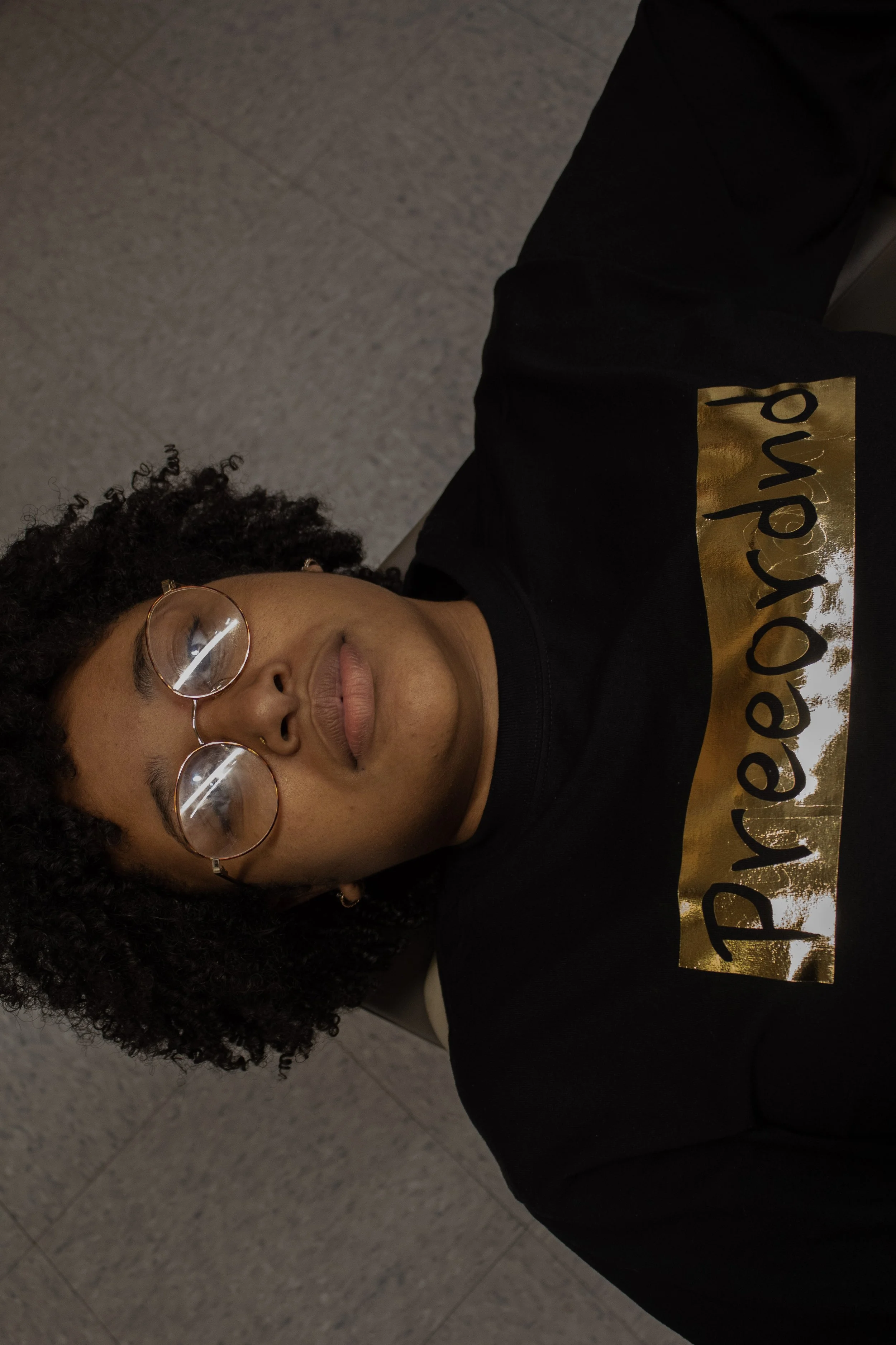 A woman with curly hair and glasses lying down, wearing a black shirt with gold company shirt.