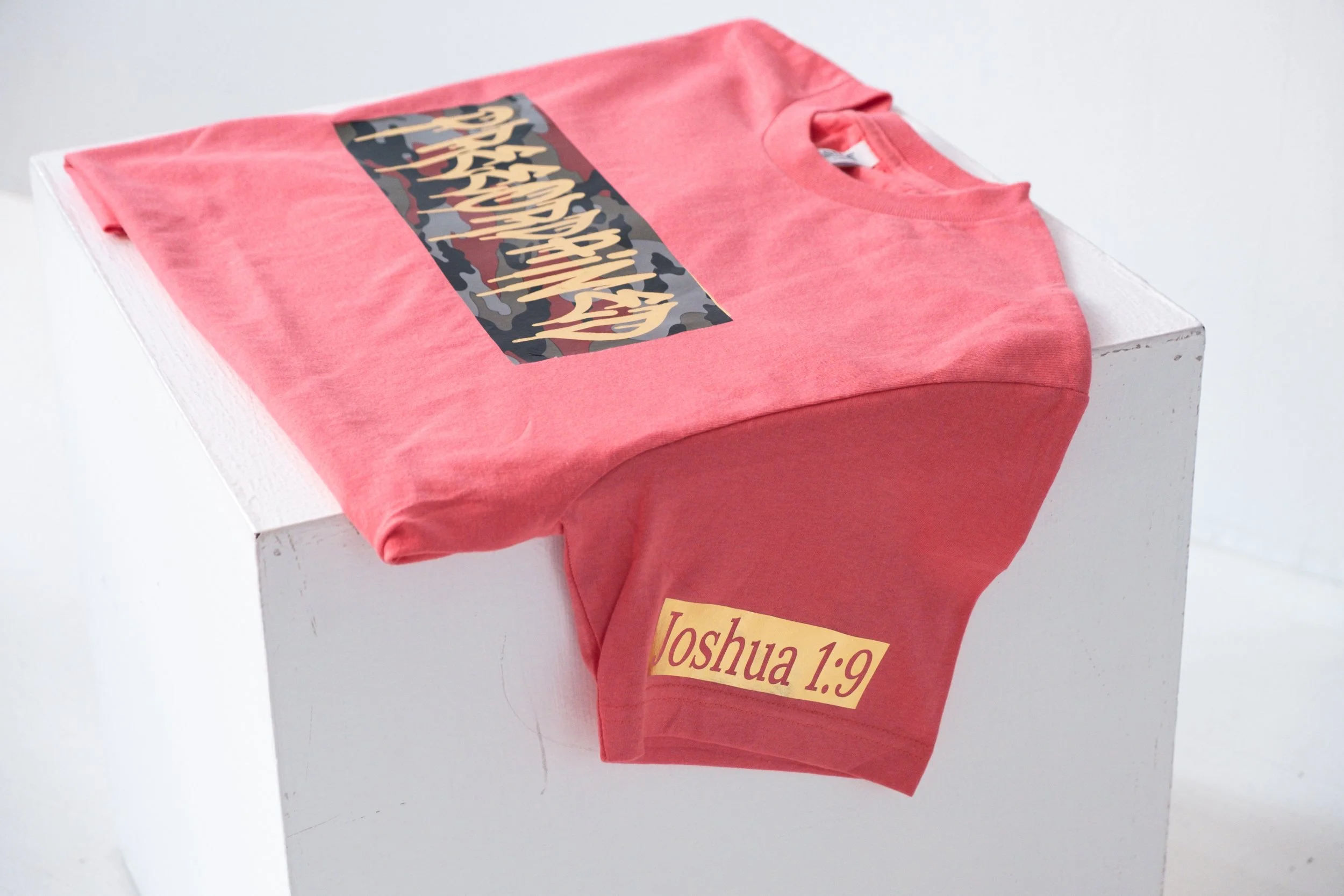 A coral color T-shirt with a red, black and gray camouflage-colored graphic and the phrase "Preeordained" written in graffiti, and a gold-platted 'Joshua 1:9' displayed on the left arm-sleeve.