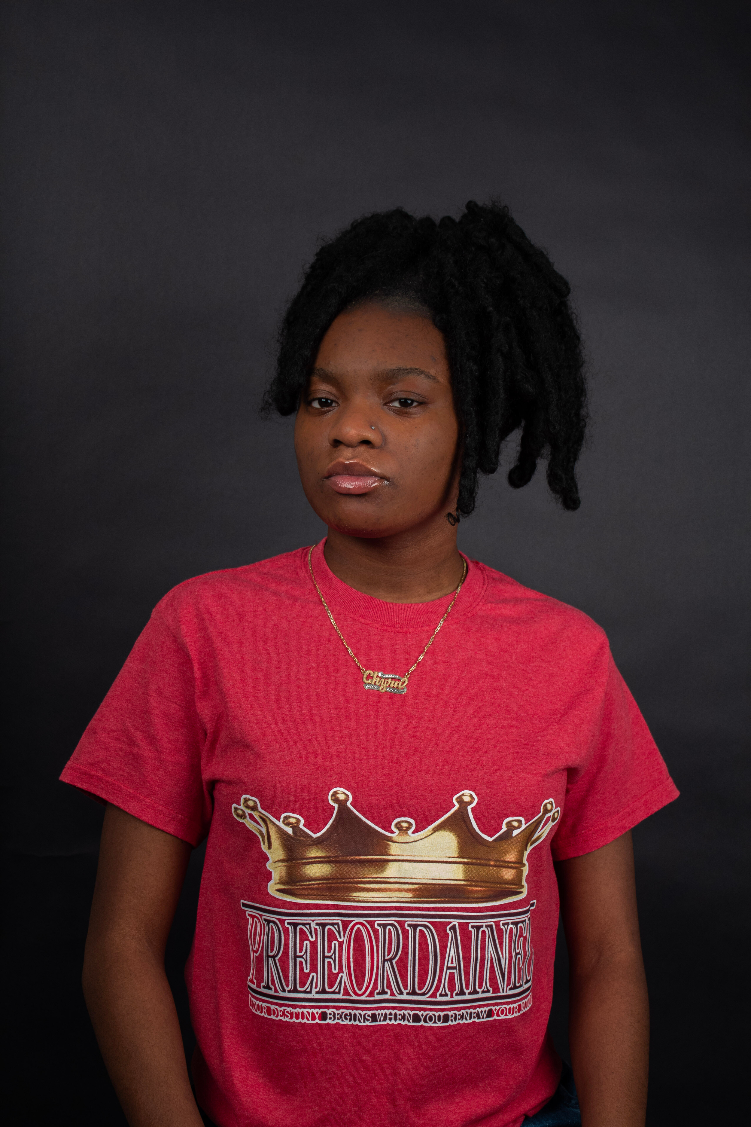 Young woman with dark curly hair, wearing a red T-shirt with a gold crown and the words 'Preordained' on it, against a dark background.