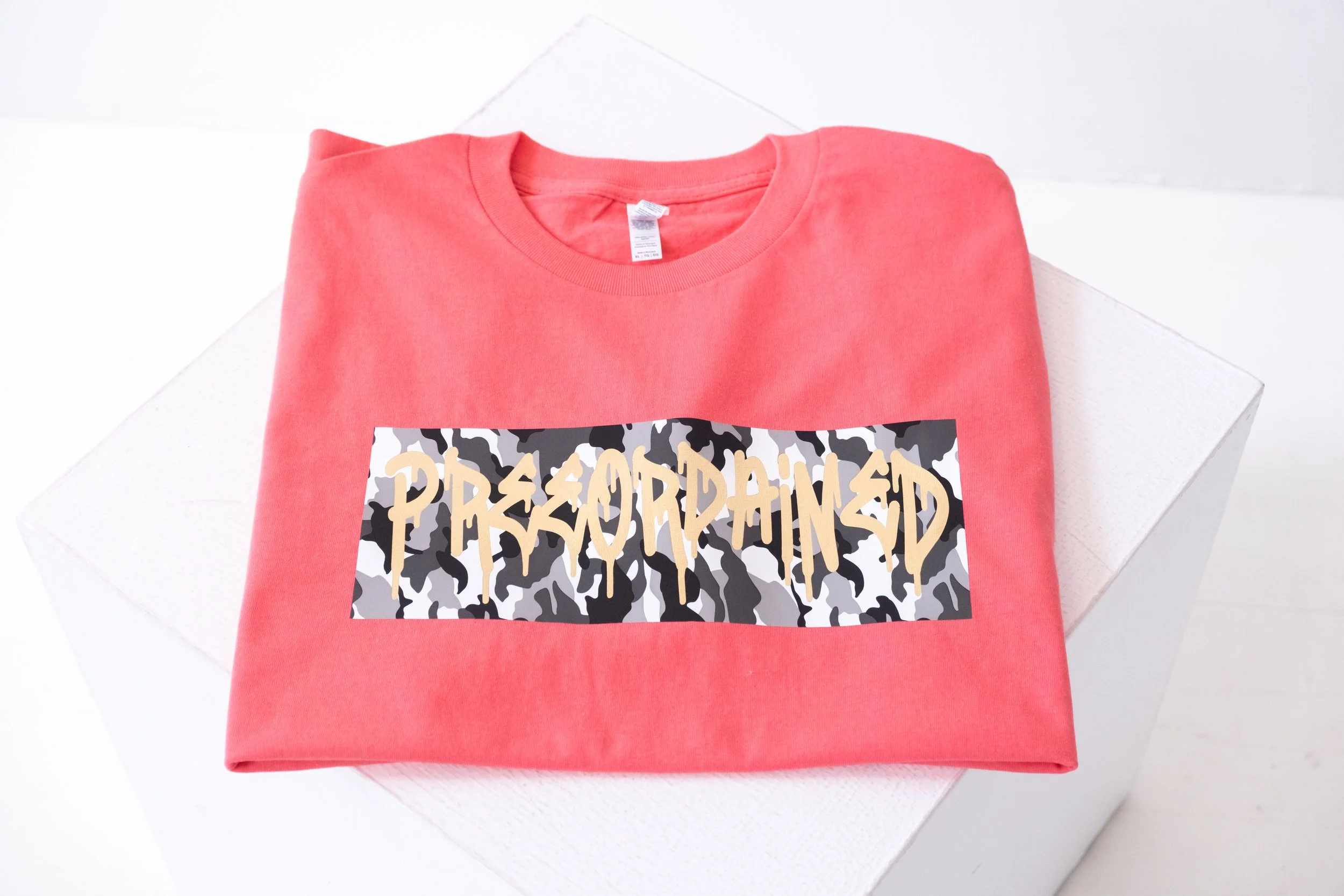 A coral color T-shirt with a white, black and gray camouflage-colored graphic and the phrase "Preeordained" written in graffiti-style font.