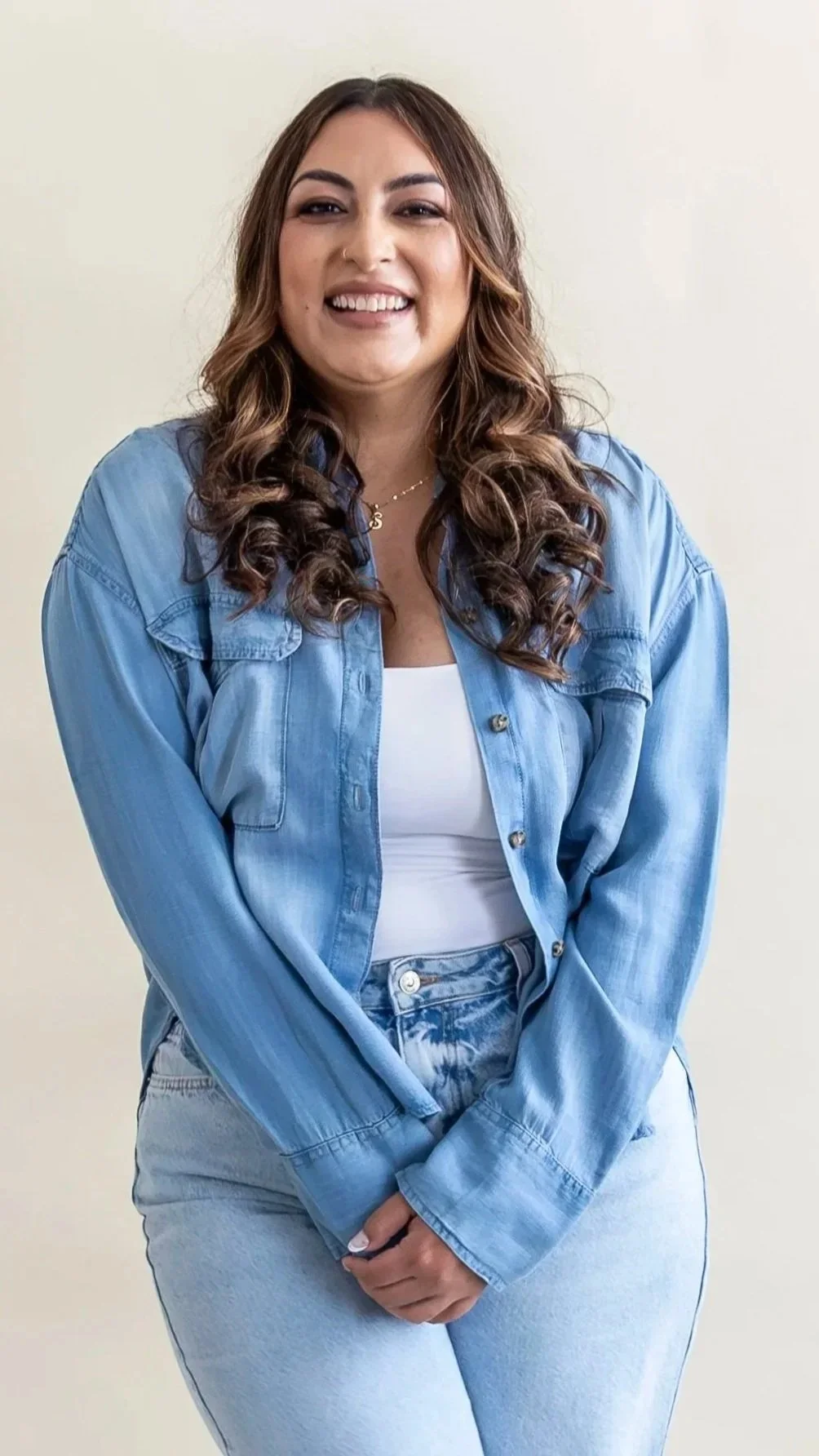 A young woman with curly brown hair, wearing a blue denim jacket over a white top, smiling at the camera against a plain light background.