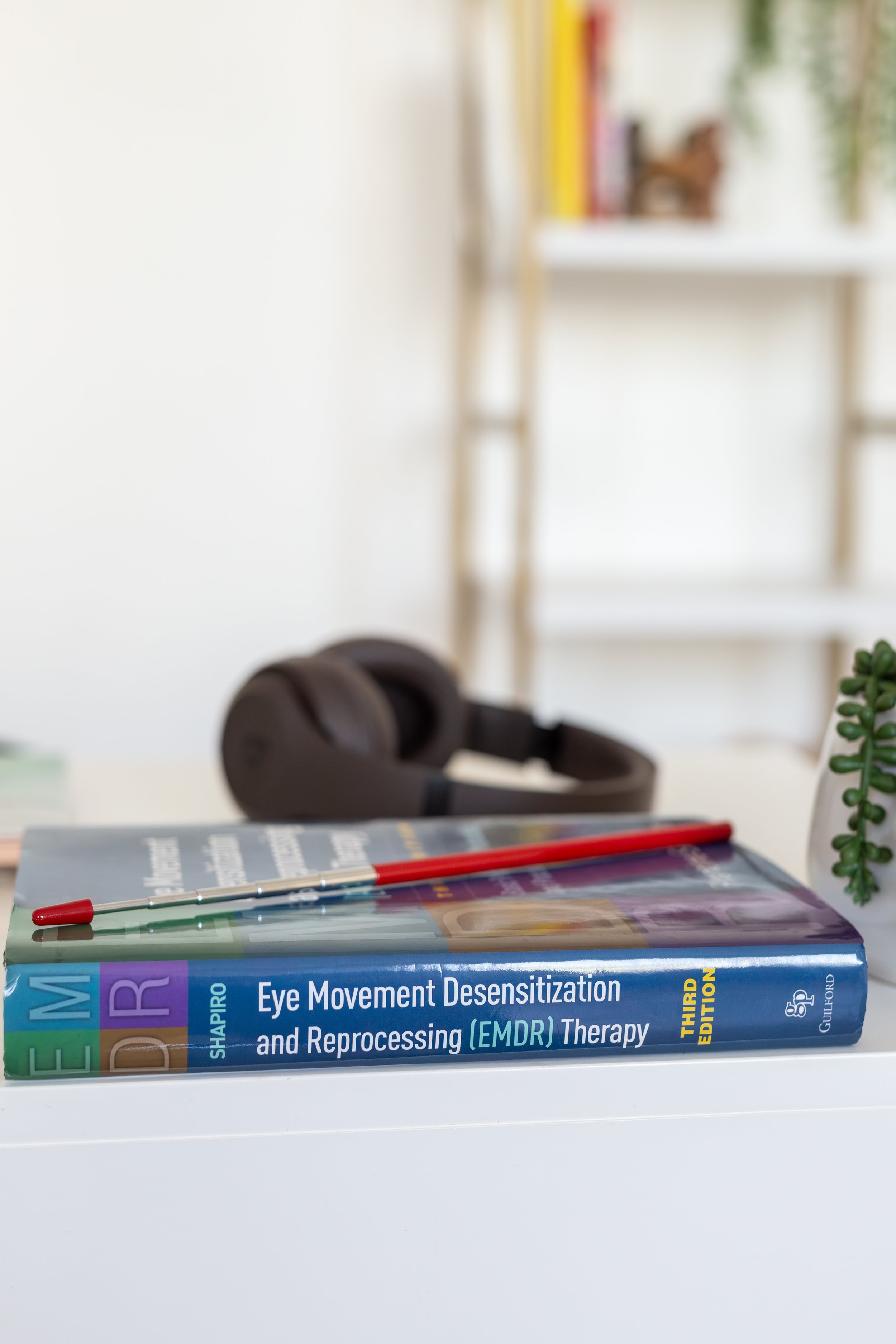 A stack of books on a white table, with a red pen on top, focusing on a book titled "Eye Movement Desensitization and Reprocessing (EMDR) Therapy." In the background, headphones, a plant, and a bookshelf are visible in a bright, modern room.