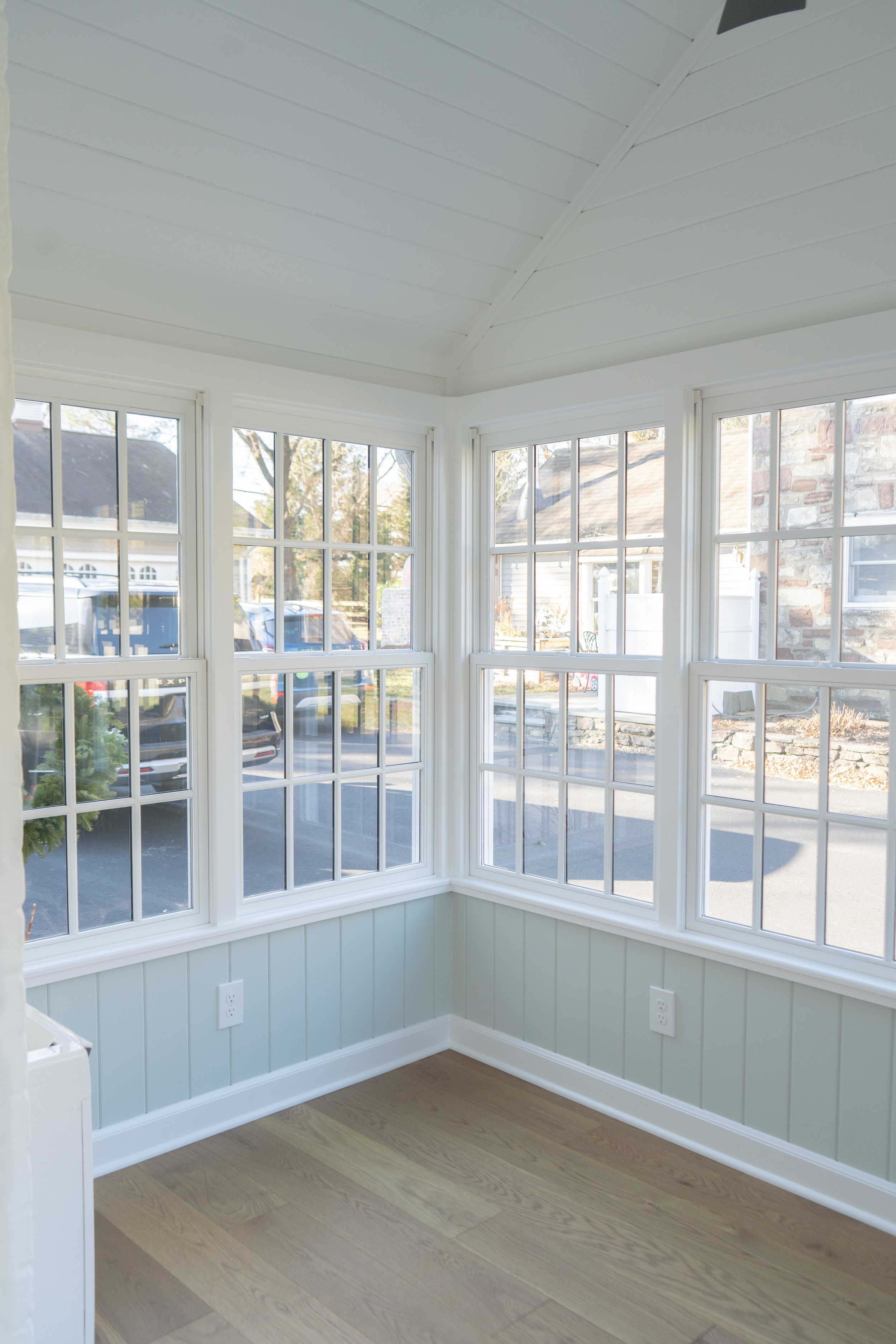 Bright sunlit corner of a room with large white-framed windows, light green beadboard wainscoting, and light wood flooring.
