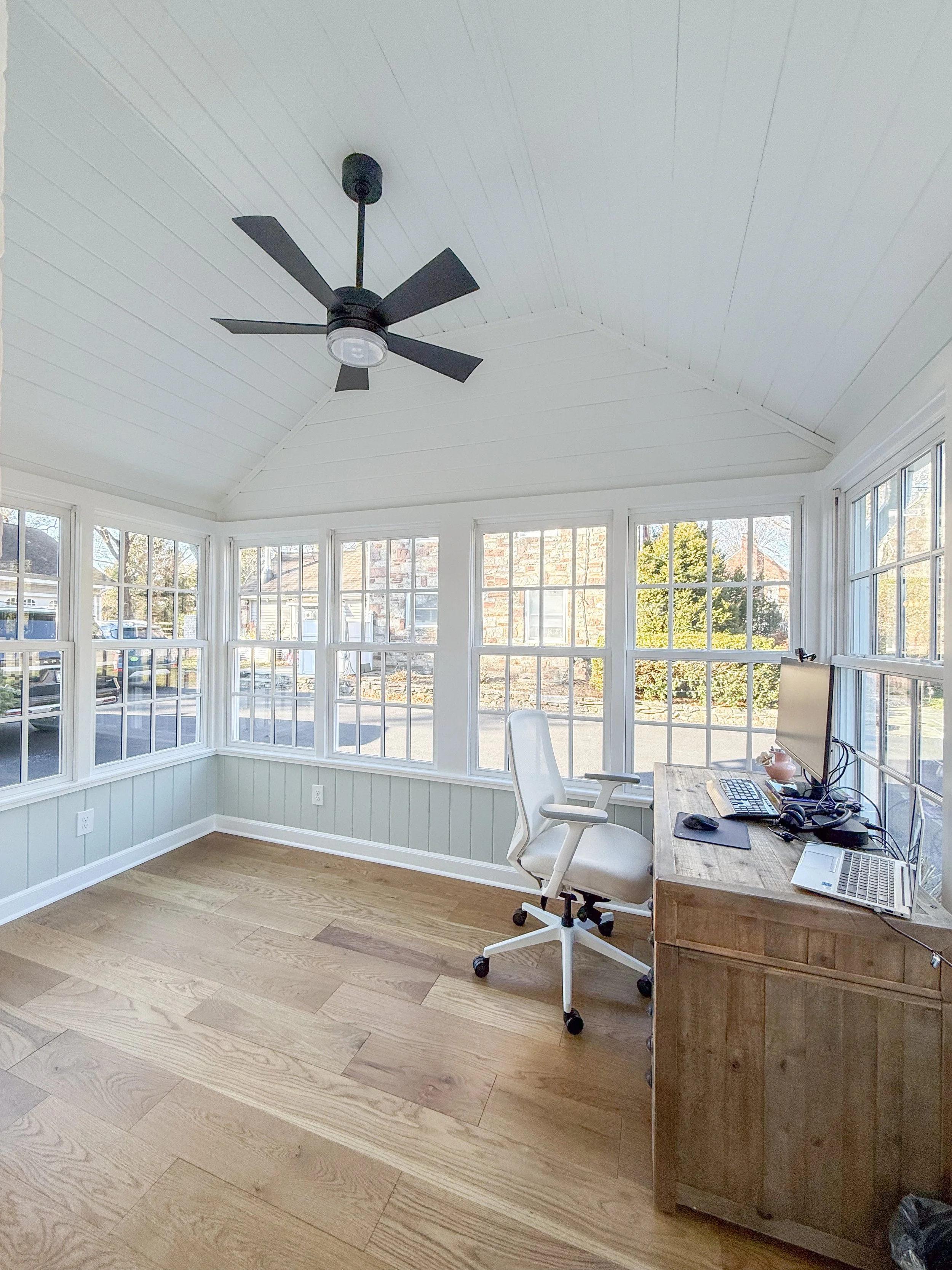 Bright sunroom with wooden floor, white walls, and large windows. Contains a desk with a computer, a white office chair, and a ceiling fan.