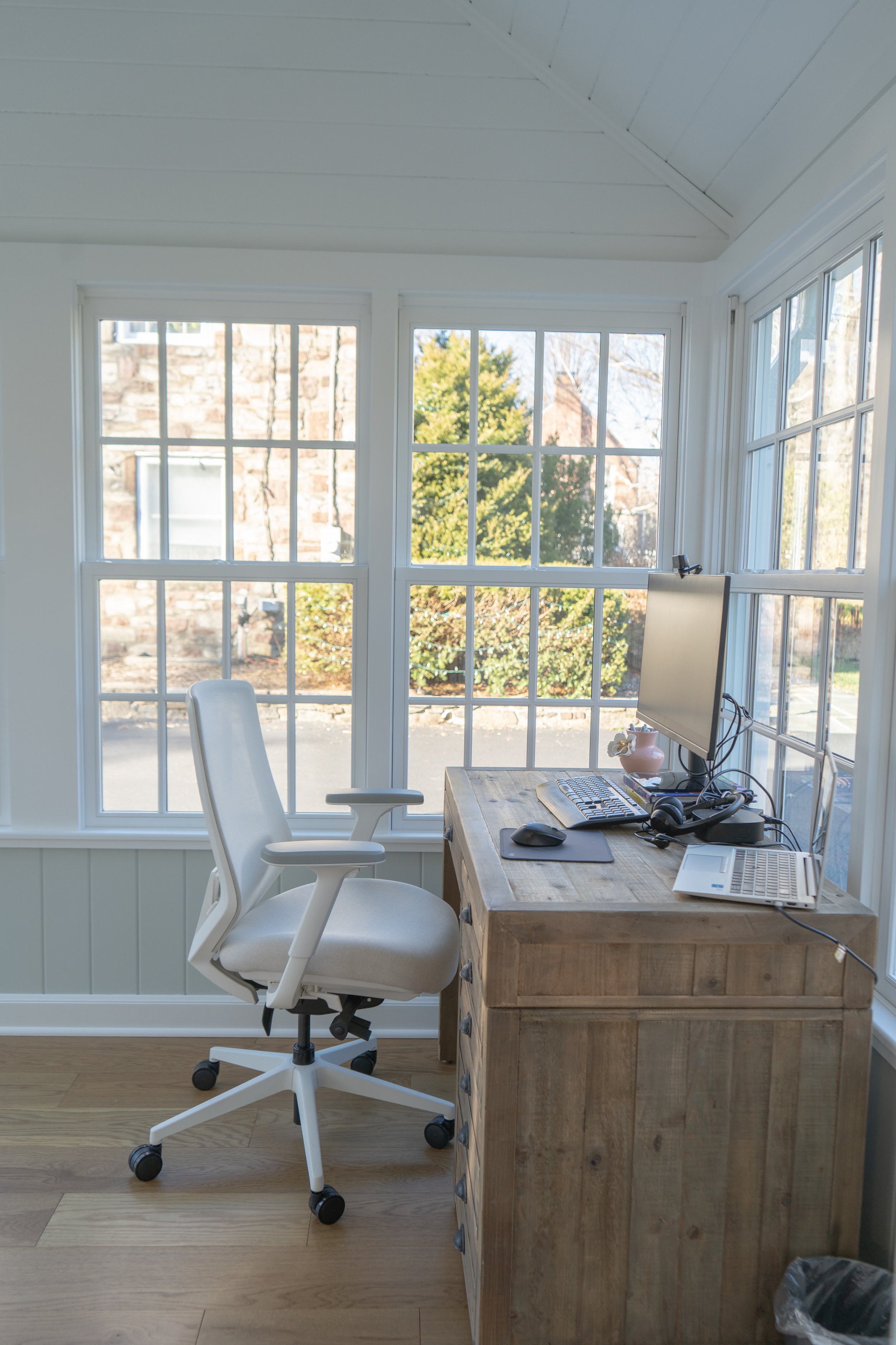 Home office with a white ergonomic chair, a wooden desk with a computer monitor, keyboard, mouse, headphones, and a small decorative vase, all set in a bright sunlit room with large windows and a view of trees and a stone wall outside.