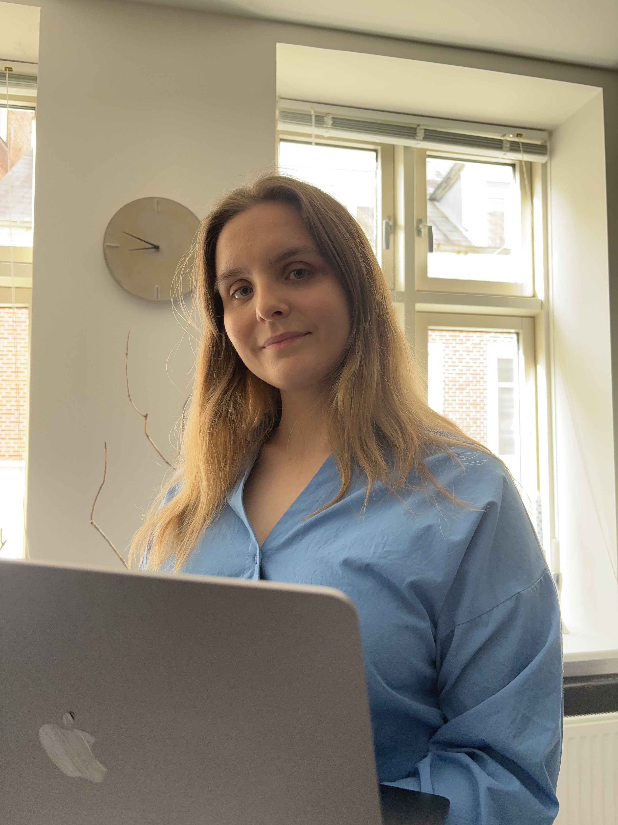 En kvinde med langt, lyst hår og blå uniform sidder foran en laptop ved et vindue med sollys.