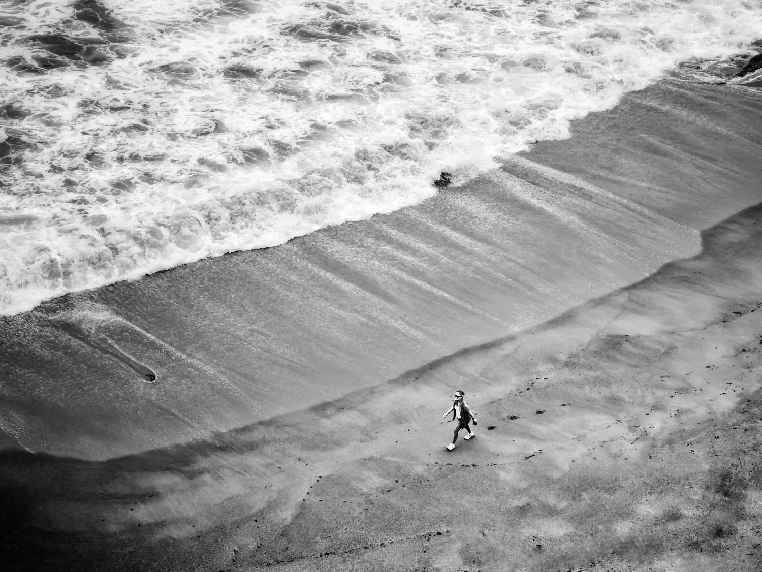 Black-and-white shoreline with a lone figure walking along textured sand.
