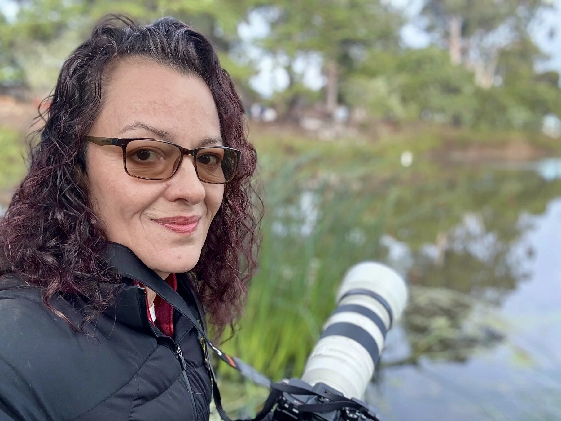 Selina smiling by a lakeside while holding a long-lens camera outdoors.