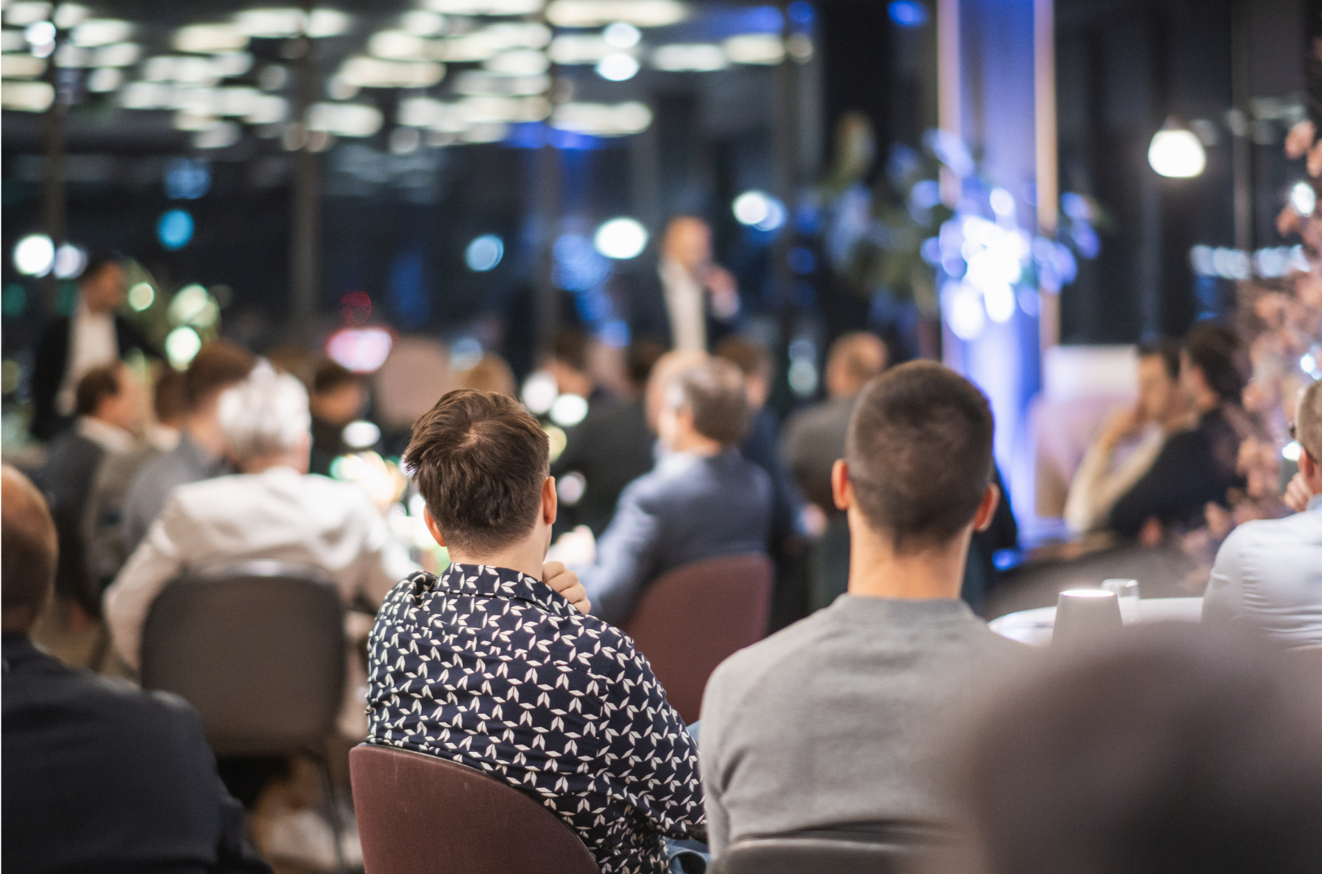 Audience seated at an indoor event with stage lights and a presenter in the background