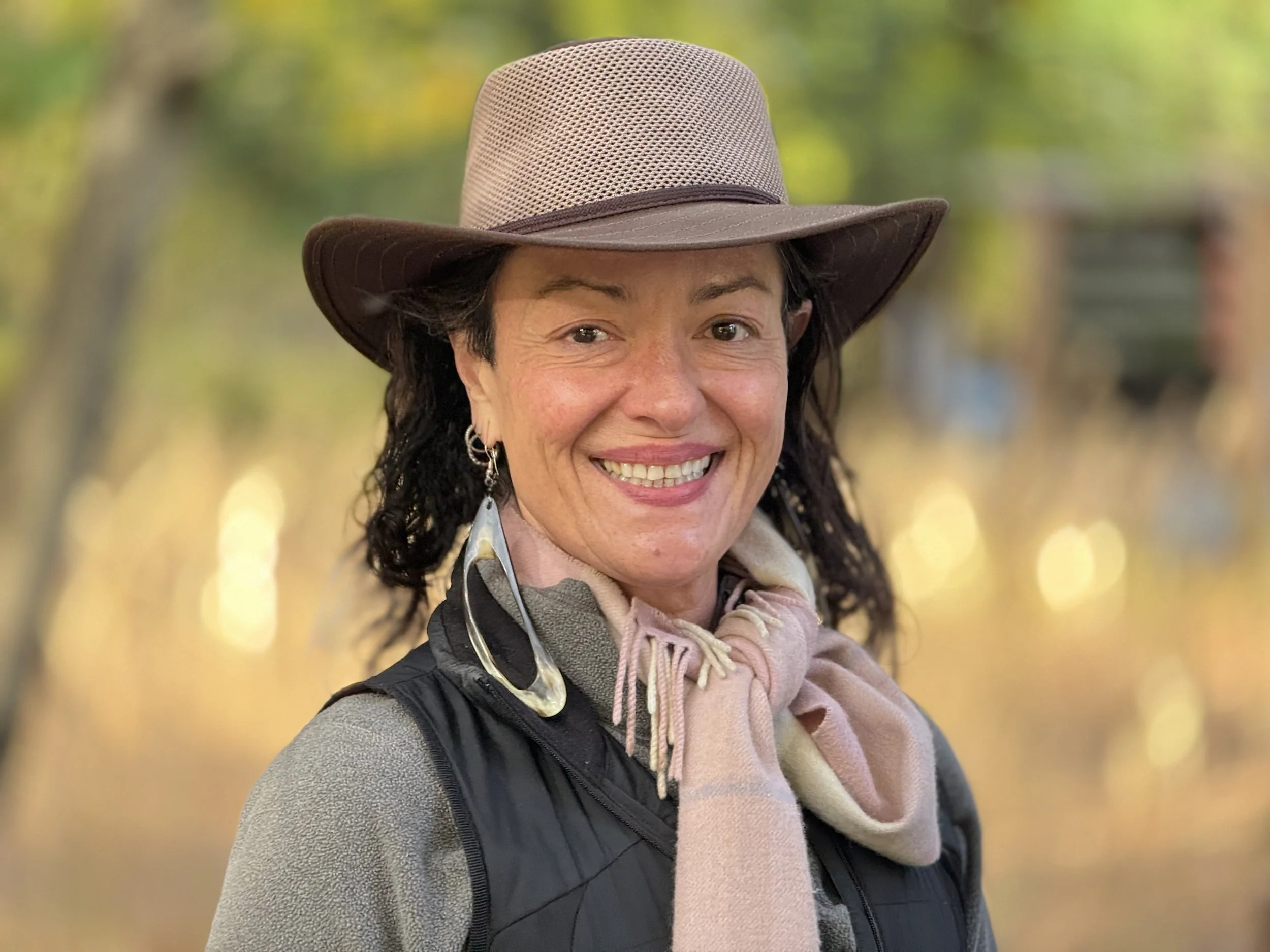 Selina smiling outdoors wearing a hat and scarf in warm natural light.