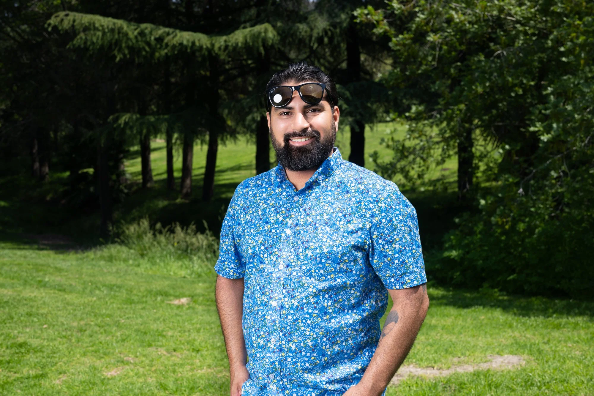 Thomas Martinez of Photos By TM in a Blue flower pattern button up short leave shirt in a field with trees in the background on set for a spring photo shoot