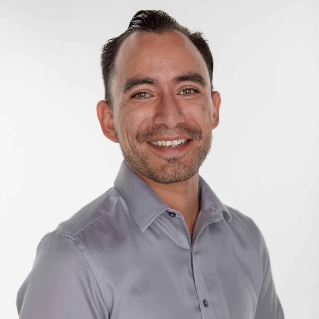Portrait of a smiling man with short dark hair, light brown eyes, and light skin, wearing a light gray button-up shirt, against a plain white background.