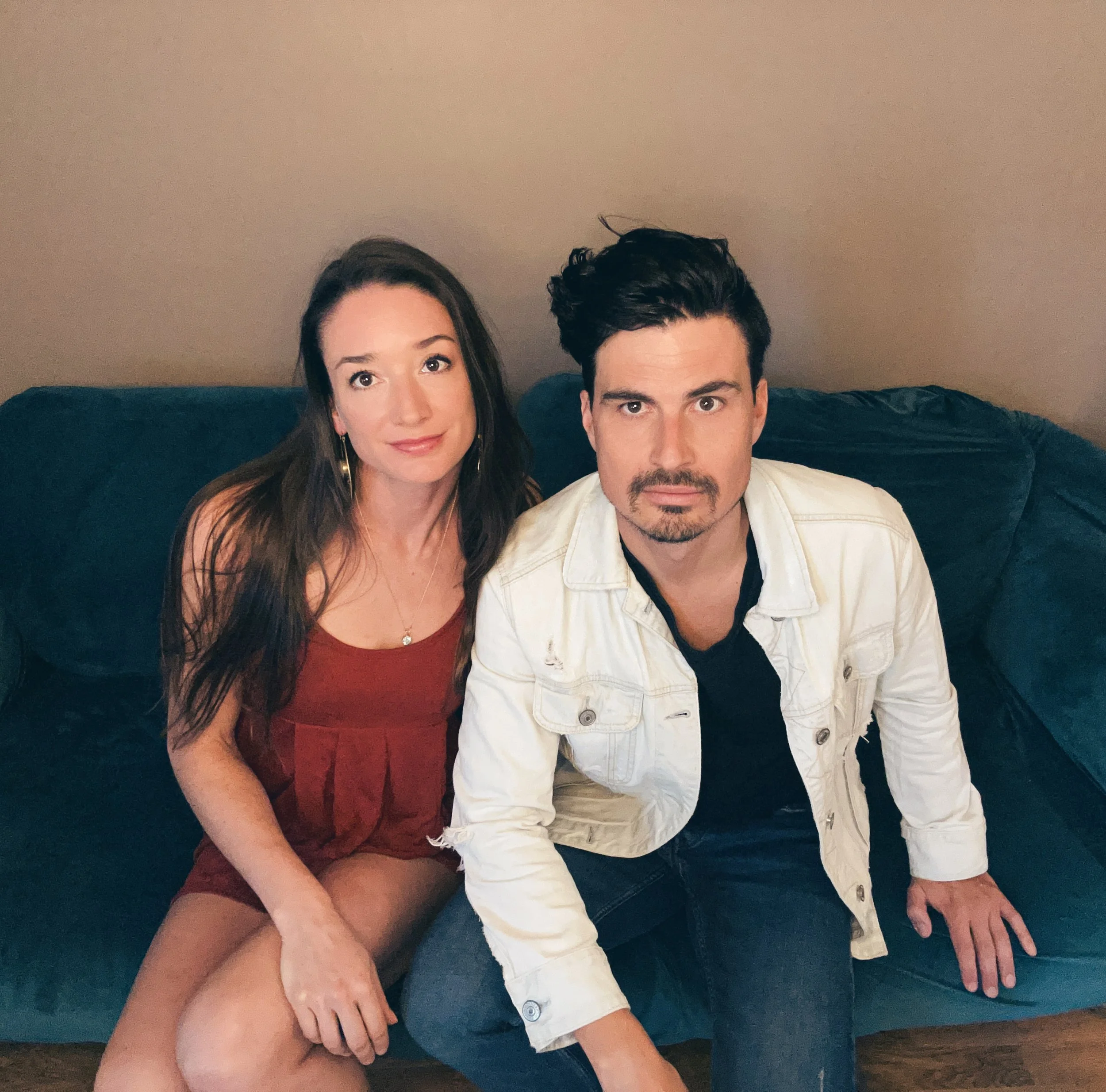 A woman in a red dress sitting next to a man in a white jacket on a teal couch against a plain wall.