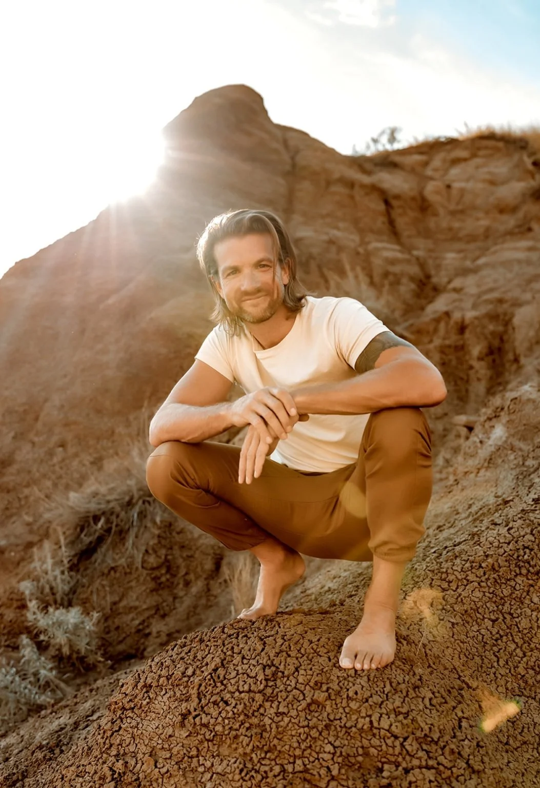 A man with long hair and a beard crouching on a rocky desert landscape during sunset, smiling at the camera.