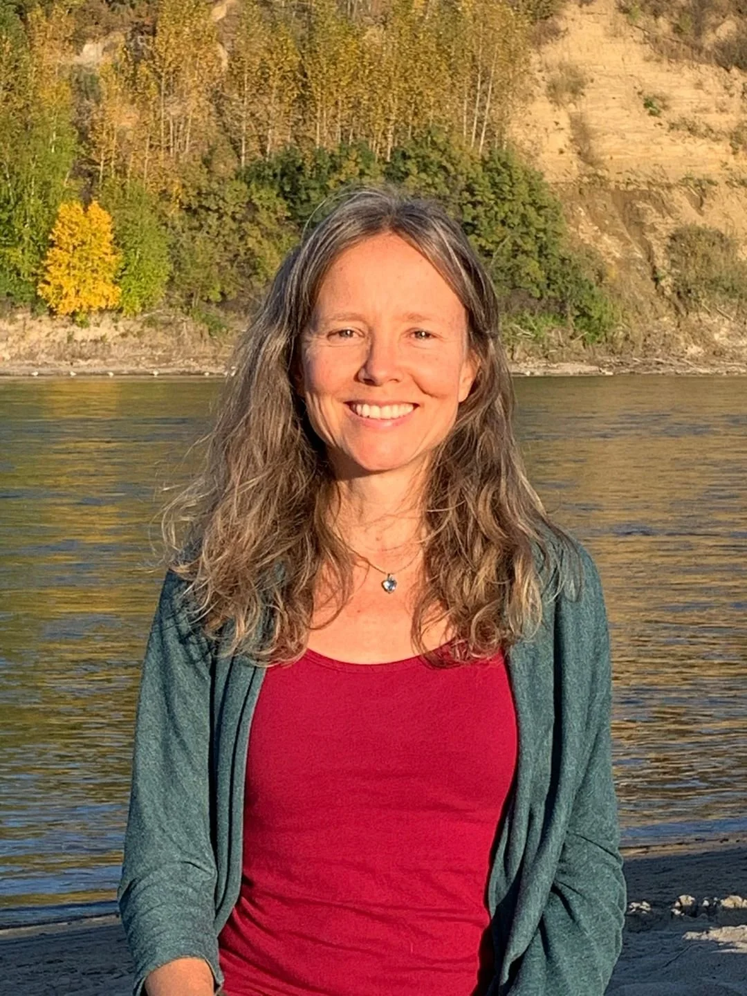 A woman with long wavy hair smiling outdoors near water with trees and a hillside in the background.