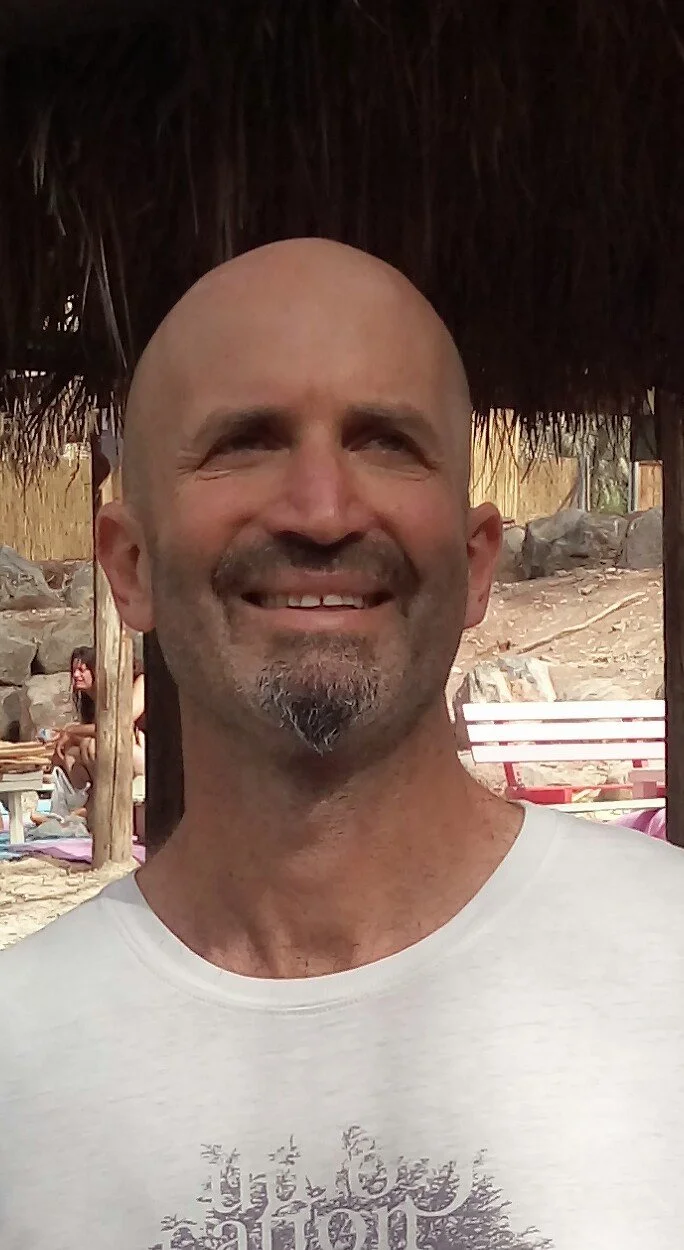 A smiling, bald man with a goatee stands outdoors with a thatched roof overhead. In the background, there are rocks, a woman sitting on a lounge chair, and a wooden bench.